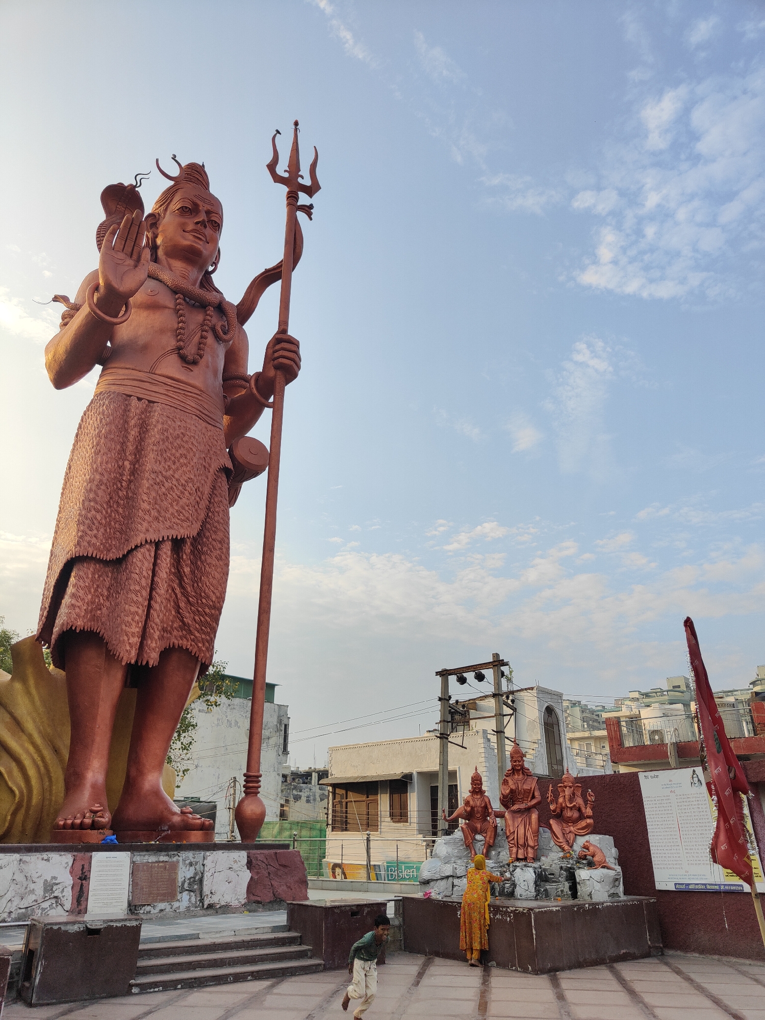 A low-angle, wide shot captures a colossal copper statue of the Hindu deity Shiva, standing prominently in Gurgaon, India. Shiva is depicted with his right hand raised in a gesture of blessing or reassurance, and he holds a trident in his left hand. The statue is adorned with a snake coiled around its neck and wrists, a common iconography associated with Shiva. The lower part of the statue is draped in a textured copper garment, resembling a skirt.
In the foreground, a young boy in a green shirt and cream-colored pants runs across a paved area. To the right of the main statue, a collection of smaller copper statues depicts other Hindu deities, including what appears to be Ganesh, seated on a rocky platform. A woman in a yellow sari is tending to these smaller statues. A red flag is visible on a pole to the far right, with text in Hindi on a sign behind it. The background reveals a mix of urban architecture, including residential buildings and what looks like electrical infrastructure. The sky above is a pale blue, with scattered white clouds. The overall scene suggests a public space, likely a temple complex or a prominent landmark in the city.