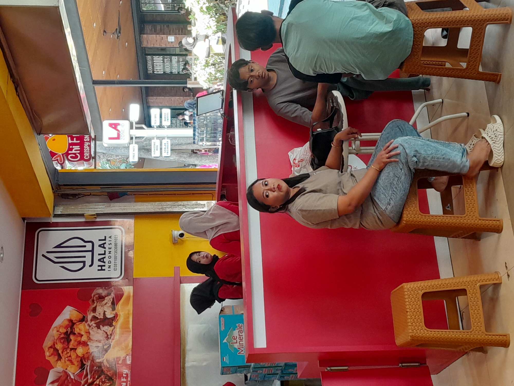 The image depicts the interior of a casual dining establishment, likely a fast-food restaurant, with a prominent red color scheme. In the background, a sign displays the "HALAL INDONESIA" logo, indicating compliance with Islamic dietary laws. To the right, a sign for "Chi Fi CRISPY SN" is visible, suggesting a chicken-based menu. Several individuals are present. Three young women, two wearing headscarves, are behind a red counter, appearing to be staff members. One woman in a red hijab looks directly at the camera with a neutral expression, while another in a black hijab looks away. A third woman, also in a red outfit and black hijab, stands between them. In the foreground, a woman in a beige t-shirt and light blue jeans sits on a brown plastic stool, facing the camera with a relaxed posture. To her right, a young man in a gray t-shirt sits on a similar stool, looking towards the foreground. Another person, partially obscured, is seated to his right. The environment is brightly lit, suggesting it's daytime. The presence of the "HALAL INDONESIA" logo and the overall atmosphere suggest this location is in Indonesia. Given the mention of Sempusari, the scene is likely within a commercial area of this city.