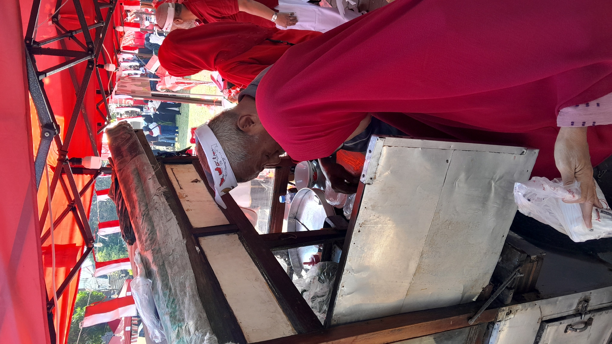 The image depicts an outdoor scene under a red and white striped canopy, likely at a market or festival. In the foreground, an elderly man wearing a white cap with a red and white map design is tending to a food cart. He appears to be preparing food, with his hands reaching into the cart. A person in a bright pink hijab is partially visible in the foreground, facing the man and the food cart. Other individuals in red clothing are also visible in the background, suggesting a communal event or gathering. The food cart itself is made of wood and metal, with various containers and bags visible inside. The overall atmosphere suggests a busy, vibrant setting where food is being served to a crowd.