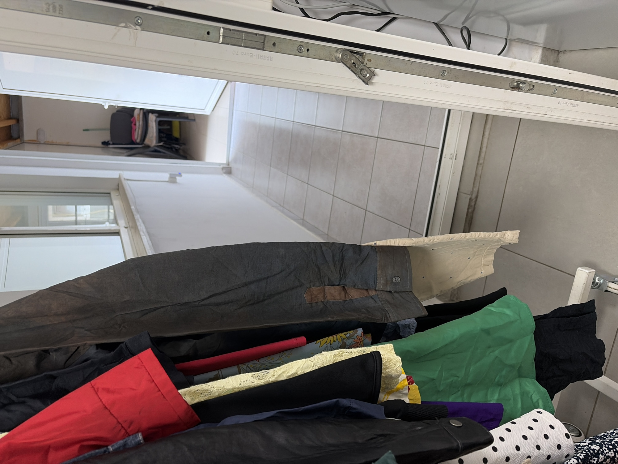 The image is a close-up, high-angle shot of a laundry rack piled with various garments. The rack appears to be positioned near an open window with white frames. A tiled floor is visible outside the window.
The clothing on the rack includes a dark gray garment with a button and a brown patch, a red item, a black garment, and a green garment. Other clothing items in various colors and patterns are also present, including a white garment with black polka dots.
Parts of the window's metal mechanism are visible. The text "RAVAL-Euro 70", "HAI-Euro 70" and "REHAL" can be seen on the window mechanism.
Due to the limited information provided, it is not possible to accurately determine the exact location within Stara Zagora, Bulgaria.