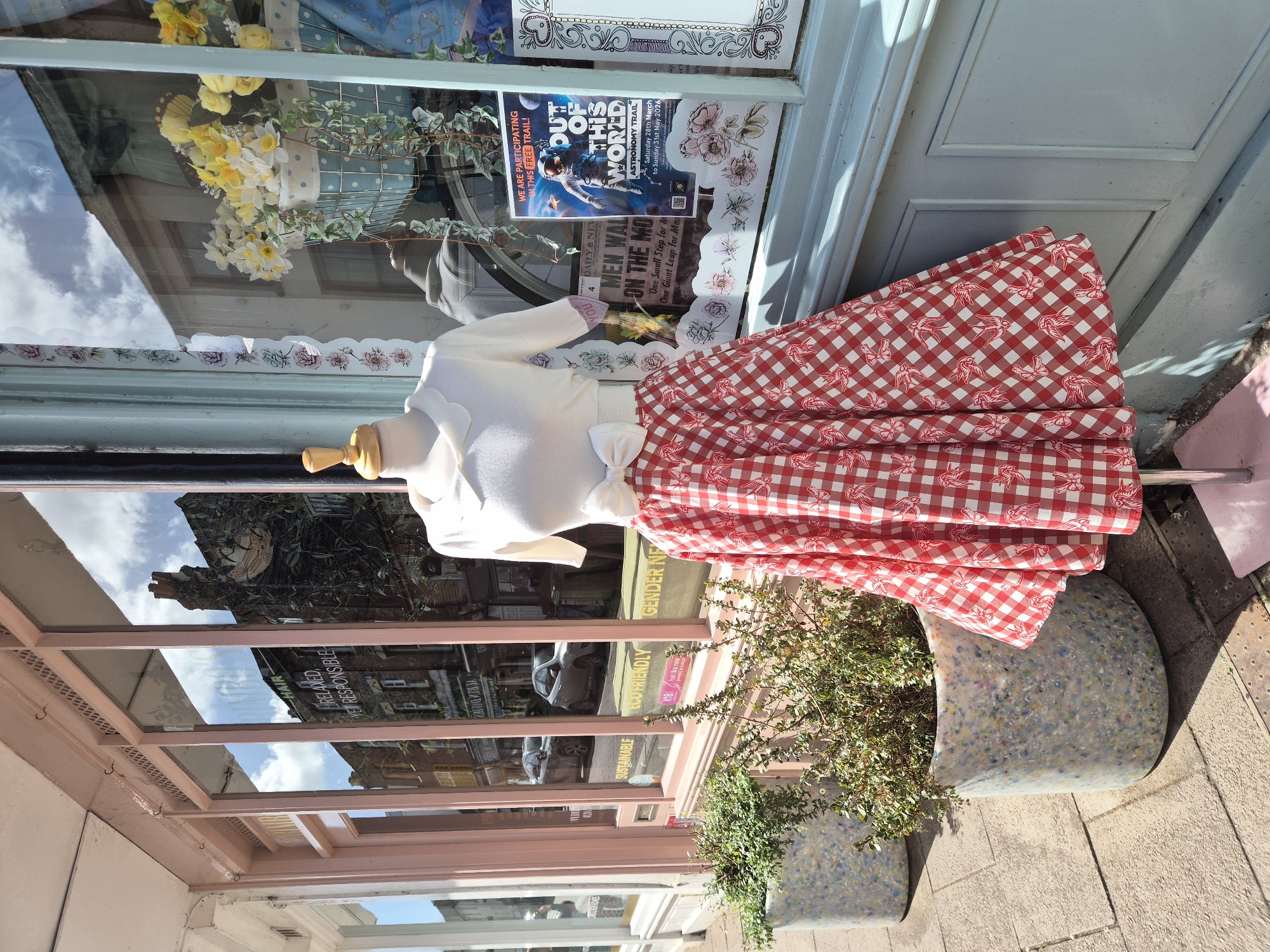 The image shows a shop display in West Suffolk, United Kingdom. A mannequin wearing a white cardigan and a red and white gingham skirt is positioned outside a shop with a light blue door. A potted plant sits next to the mannequin. The shop window reflects the street and buildings opposite. Posters are visible inside the window, one advertising an "Out of This World Astronomy Trail" running from March to May 2026. Text such as "HAIR," "RELAXED YET RESPONSIBLE," "SUSTAINABLE," and "ECOFRIENDLY GENDER INF" are readable on a sign within the shop's reflection. A newspaper clipping about the moon landing is also visible.