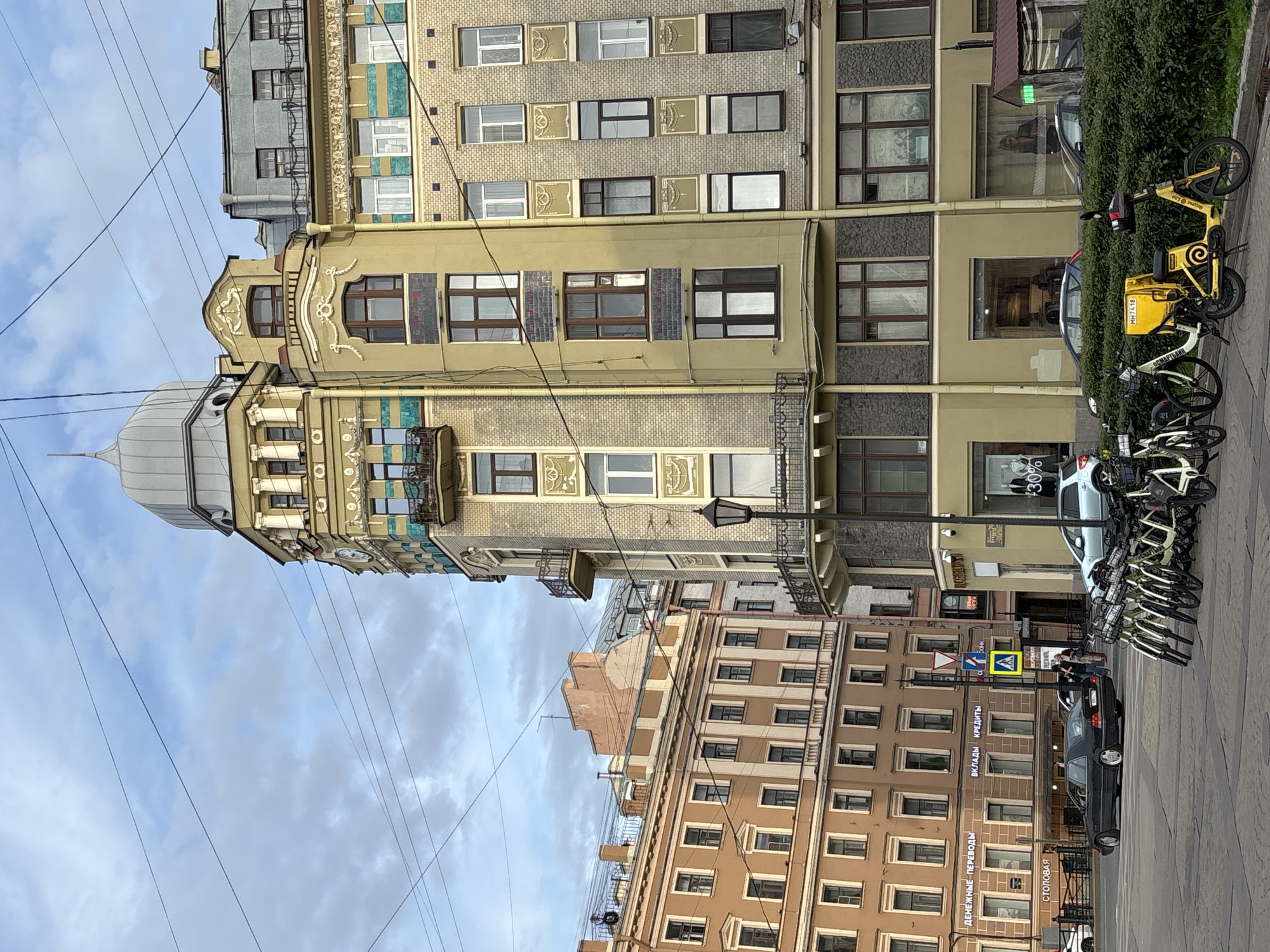 This daytime image captures an urban street scene in Saint Petersburg, Russia, under a cloudy sky. The main subject is a tall, ornate building with a distinctive light-colored facade featuring intricate architectural details, balconies, and a domed top with a spire. Adjacent to it is a building with a beige, brick-patterned facade adorned with decorative panels around its windows. Further down, a multi-story brown/tan building with numerous rectangular windows lines the street.
The street level shows a mix of activity. Several vehicles are present, including a black sedan whose license plate partially reveals the "78" region code (for Saint Petersburg) and a light-colored car partially obscured. A prominent feature is a line of parked bicycles, some of which are bright yellow bike-sharing bikes, one with a delivery box displaying "МЧ-7418". These bikes are parked next to a green hedge separating the sidewalk from a commercial frontage. A lamppost and overhead power lines crisscross the sky.
Street signs, including a pedestrian crossing sign and a yellow diamond sign (likely a main road sign), are visible. Storefronts on the brown building display text in Russian, including "ВКЛАДЫ КРЕДИТЫ" (Deposits, Loans), "ДЕНЕЖНЫЕ ПЕРЕВОДЫ" (Money Transfers), and "СТОЛОВАЯ" (Canteen/Dining Hall). A person's reflection can be seen in a shop window. The scene depicts a typical busy city environment with historical architecture and modern urban elements.