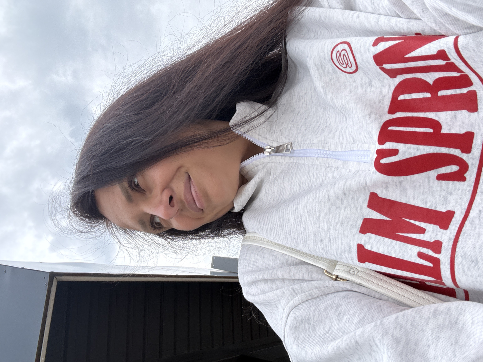 The image is a selfie of a woman, taken from a low angle and rotated 90 degrees clockwise. When viewed upright, a young woman with long, dark brown hair looks upwards into the camera with a subtle, friendly smirk. She is wearing a light grey hooded sweatshirt with a zipper closure. A light-colored strap, possibly from a shoulder bag or purse, is visible over her right shoulder, tucked behind the hoodie.
The front of her sweatshirt features large, bold red letters. Portions of the visible text read "SPRIN" and "M", strongly suggesting the phrase "PALM SPRING". Above this text, a small circular red logo, possibly a stylized 'S', is also present.
The background consists primarily of a cloudy, overcast sky, which appears light grey. Below her, part of a dark, angular building or structure with paneling is visible, suggesting an outdoor setting. The cloudy weather provides soft, diffused lighting. The location context provided is Chaikovsky, Russia.
