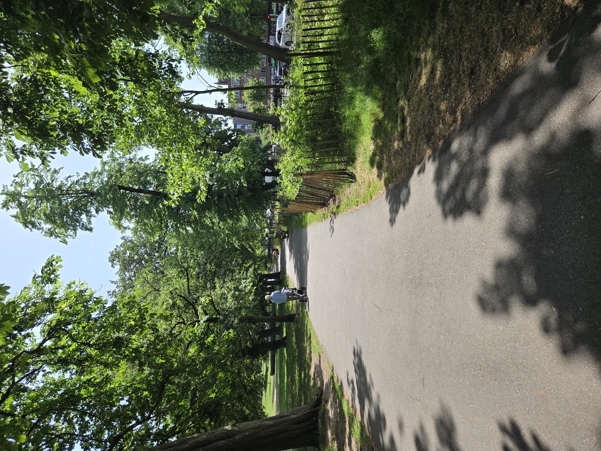 A straight asphalt pathway lined with trees and a thin wooden fence stretches into the distance. A person in a light blue shirt is walking along the path. Buildings and parked cars are visible in the background. The location is likely within a park in London, United Kingdom.