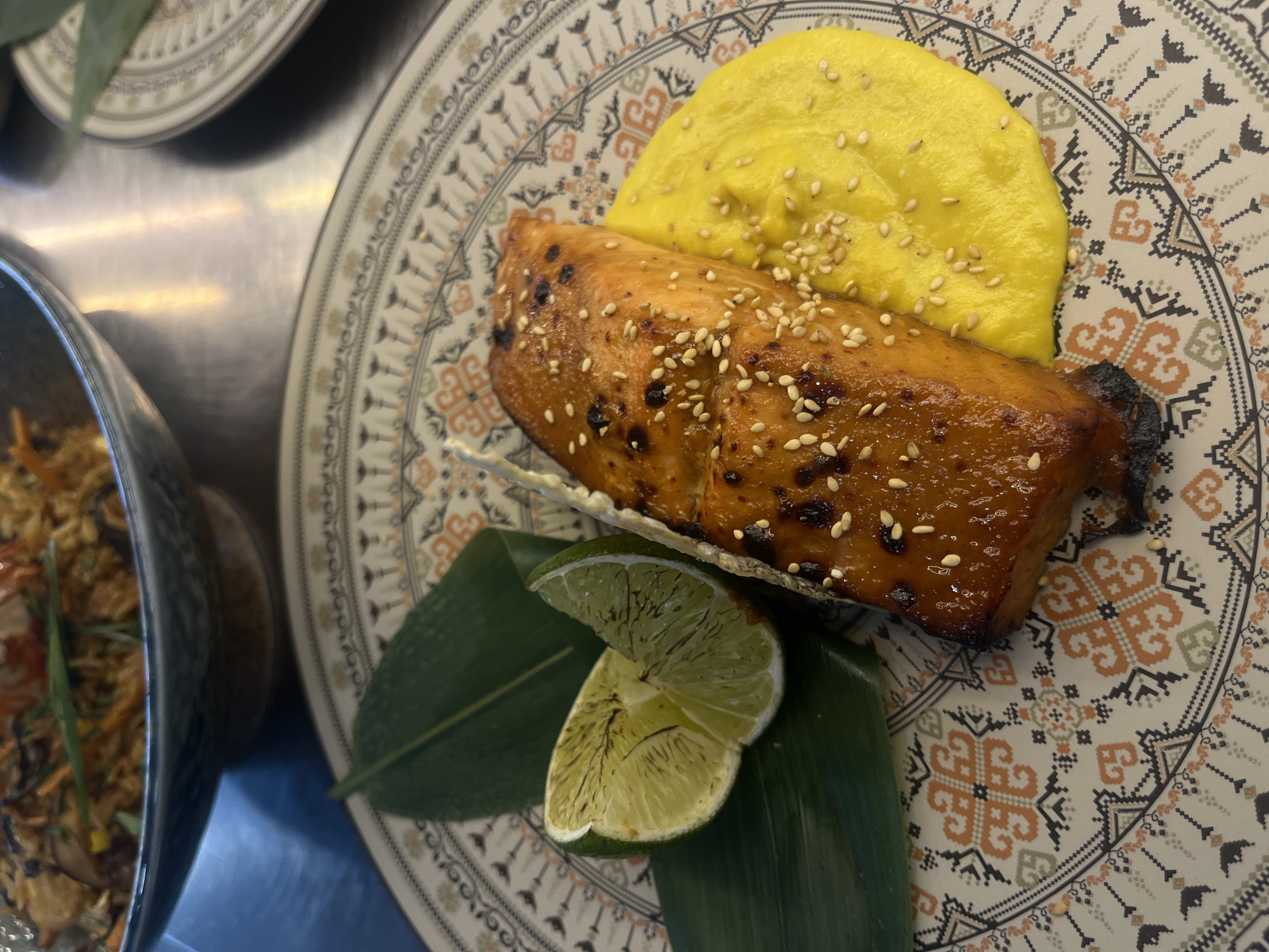 The image shows a plate of food, likely taken in Sofia, Bulgaria. A piece of glazed fish sprinkled with sesame seeds rests on a green leaf alongside two lime slices. A dollop of yellow purée, also sprinkled with sesame seeds, sits beside the fish. The plate has an intricate, patterned design in beige, orange, and gray. To the left, partially visible, is a bowl containing a dish, possibly rice or noodles, with mixed vegetables. Another plate is visible in the background. The setting appears to be a restaurant or dining environment, possibly in Sofia.