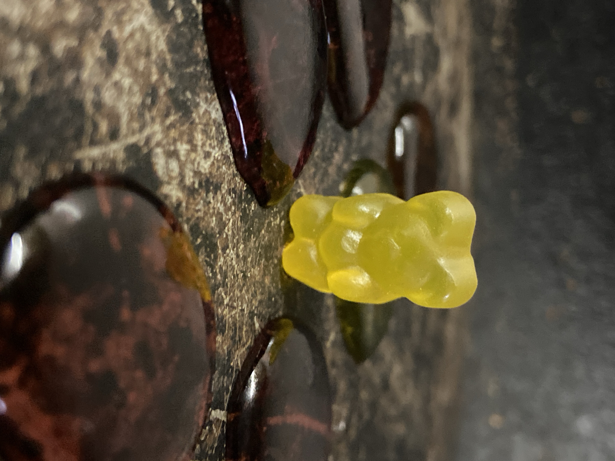 A close-up, high-angle shot captures a still life arrangement featuring a single yellow gummy bear and several dark, glossy objects on a textured, mottled surface. The yellow gummy bear is the central focus, positioned horizontally slightly to the right of the image's vertical center. It appears translucent and somewhat stretched or irregularly shaped.
Surrounding the gummy bear are multiple dark, irregularly shaped, glossy objects, likely decorative glass beads or polished stones, with colors ranging from deep reddish-brown to dark green. One large, out-of-focus dark object occupies the bottom left corner, while other smaller ones are partially visible in the top left, top right, and directly behind the gummy bear. These objects reflect light, creating bright highlights on their surfaces.
The underlying surface is dark and textured, resembling a stone countertop or a rough, patterned material. The lighting appears to be artificial, highlighting the translucency of the gummy bear and the reflective quality of the dark objects. The depth of field is shallow, with the gummy bear and immediate surroundings in sharp focus, while the foreground and background elements are blurred. There is no visible text, and no specific time of day, weather, or emotions are discernible from the image.