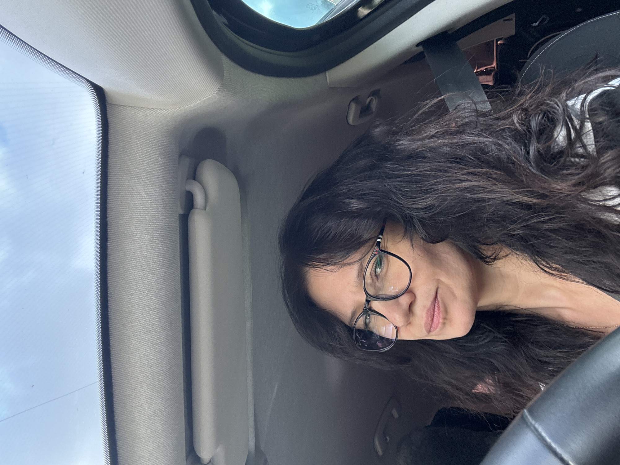 A woman is seated in the driver's seat of a vehicle, looking towards the camera. She has long, dark, wavy hair and is wearing large, round, black-rimmed glasses. Her lips are slightly parted, and she has a neutral expression. She is wearing a dark top with a white patterned layer underneath, and a gold necklace. The interior of the car is visible, including the steering wheel in the foreground, the roof lining, a sun visor, and a side door frame with a window. The sky is visible through the windshield and sunroof. The image appears to be taken during the daytime, given the bright lighting. The location is not specified as Chaikovsky, Russia.