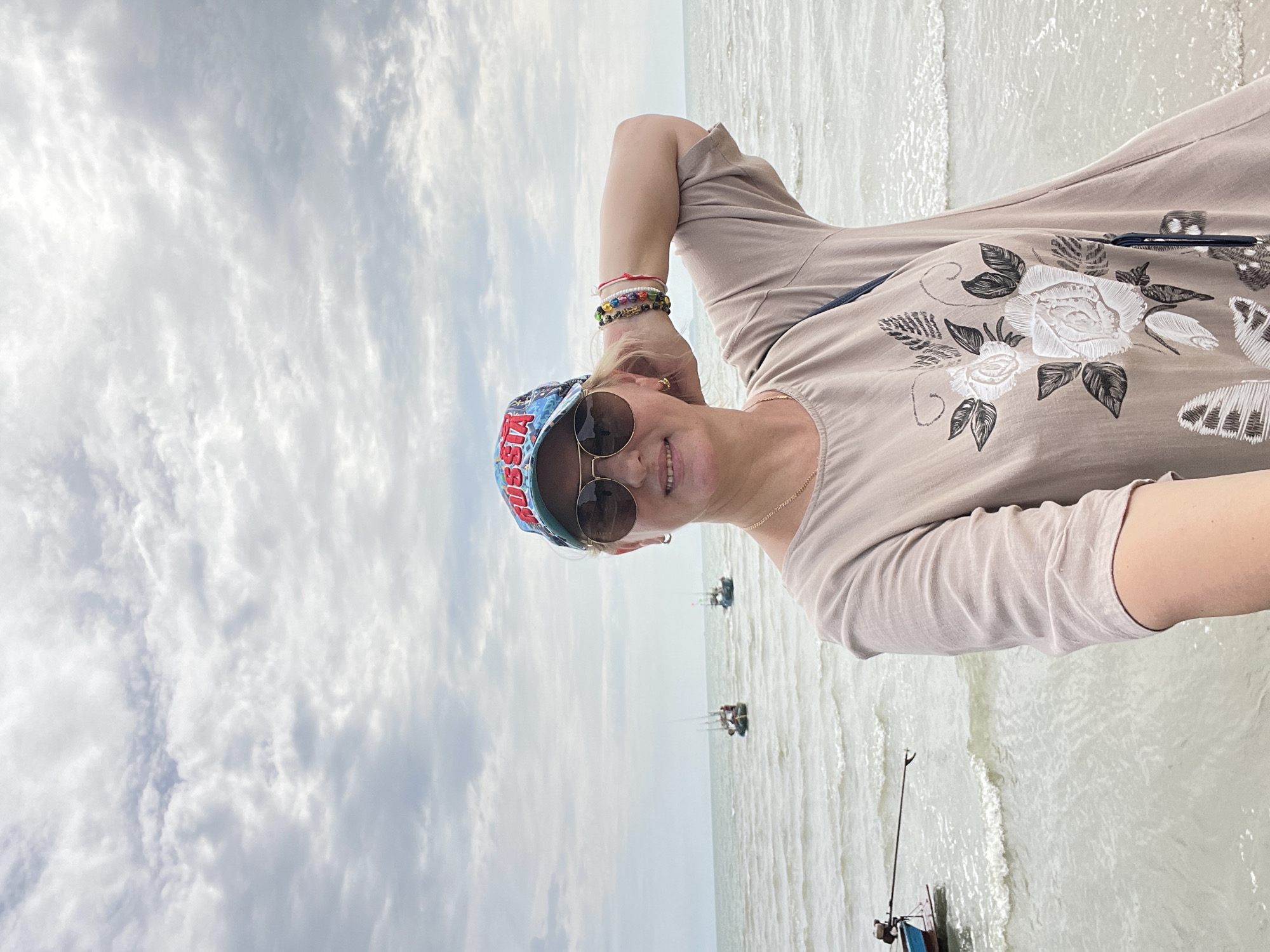 A fair-skinned woman with blonde hair and sunglasses smiles at the camera while standing on a pier overlooking the ocean. She is wearing a light brown, loose-fitting dress with a floral and butterfly print, a blue baseball cap with "RUSSIA" in red letters, and several beaded bracelets on her right wrist. Her right arm is bent at the elbow, with her hand resting behind her head. The sea behind her is calm with small waves, and several small fishing boats are visible in the distance. The sky is overcast with scattered clouds. This scene is consistent with the coastal areas of Pattaya City, Thailand.