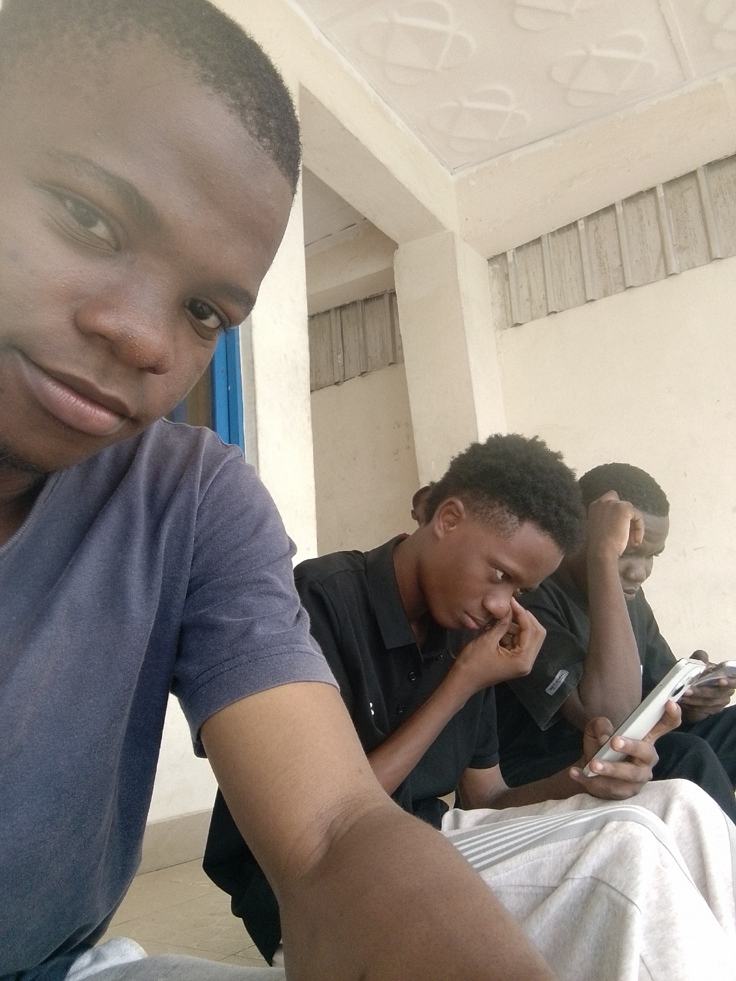 The image shows three young men sitting together in an outdoor area. The foreground figure, facing the camera, wears a dark blue T-shirt. Two other men are seated behind him. One is wearing a black collared shirt and appears to be picking at his nose. The other man is wearing a black shirt and holding a white cell phone. They are all seated on a tiled surface, with a wall visible in the background. The setting seems to be a simple outdoor area with a tiled ceiling. The location is Ife, Nigeria. The men appear to be in their late teens or early twenties.