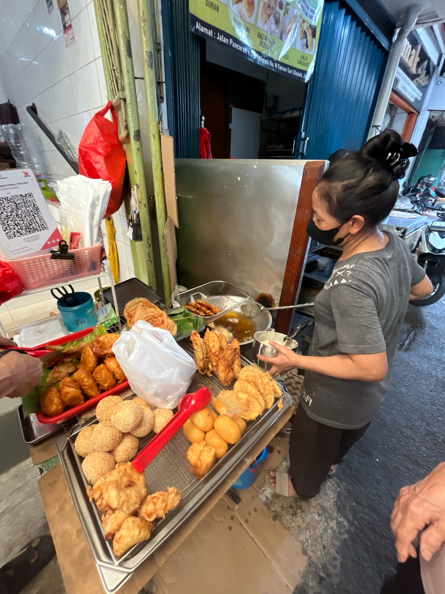 A woman wearing a black face mask and a grey t-shirt stands at an outdoor food stall in an urban environment. She is holding a metal cup and a metal utensil, seemingly in the process of preparing food. The stall displays a variety of fried food items on metal trays, including what appear to be fried bananas and sesame seed-covered pastries. Behind her, a deep fryer is visible, along with cooking utensils. A red plastic bag hangs from a green metal pole to the left of the stall. The ground is wet, indicating recent rain or cleaning. In the background, a blue corrugated metal shutter is partially visible, and a sign with text in a foreign language is mounted above it. A motorcycle is parked to the right of the stall. The scene suggests a busy street food operation.