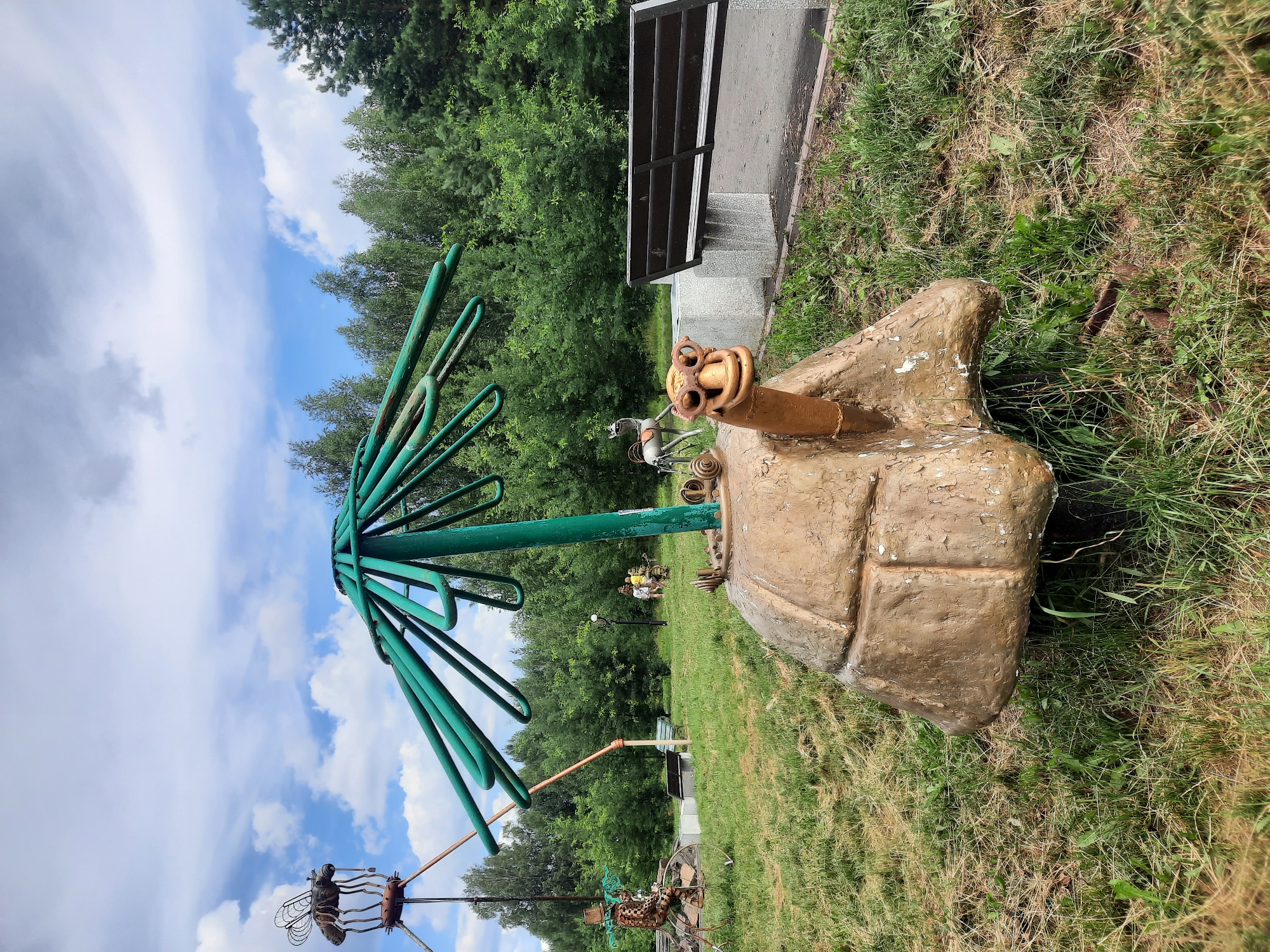 In the city of Zelenogorsk, Russia, on a grassy lawn under a cloudy sky, a whimsical metal sculpture of a turtle wearing oversized goggles sits prominently. Its shell is rough and tan, resembling a large rock, and a green, umbrella-like structure emerges from its back. To the left, another metal sculpture of a mosquito-like creature perches atop a tall, rusty pole. Further in the background, a metal sculpture of a person riding a bicycle is visible, along with other abstract metal artworks. A wooden park bench sits to the right of the turtle sculpture, positioned near a paved pathway that leads towards a dense forest bordering the area. Several people are scattered throughout the park, appearing to observe the sculptures and enjoy the outdoor space.