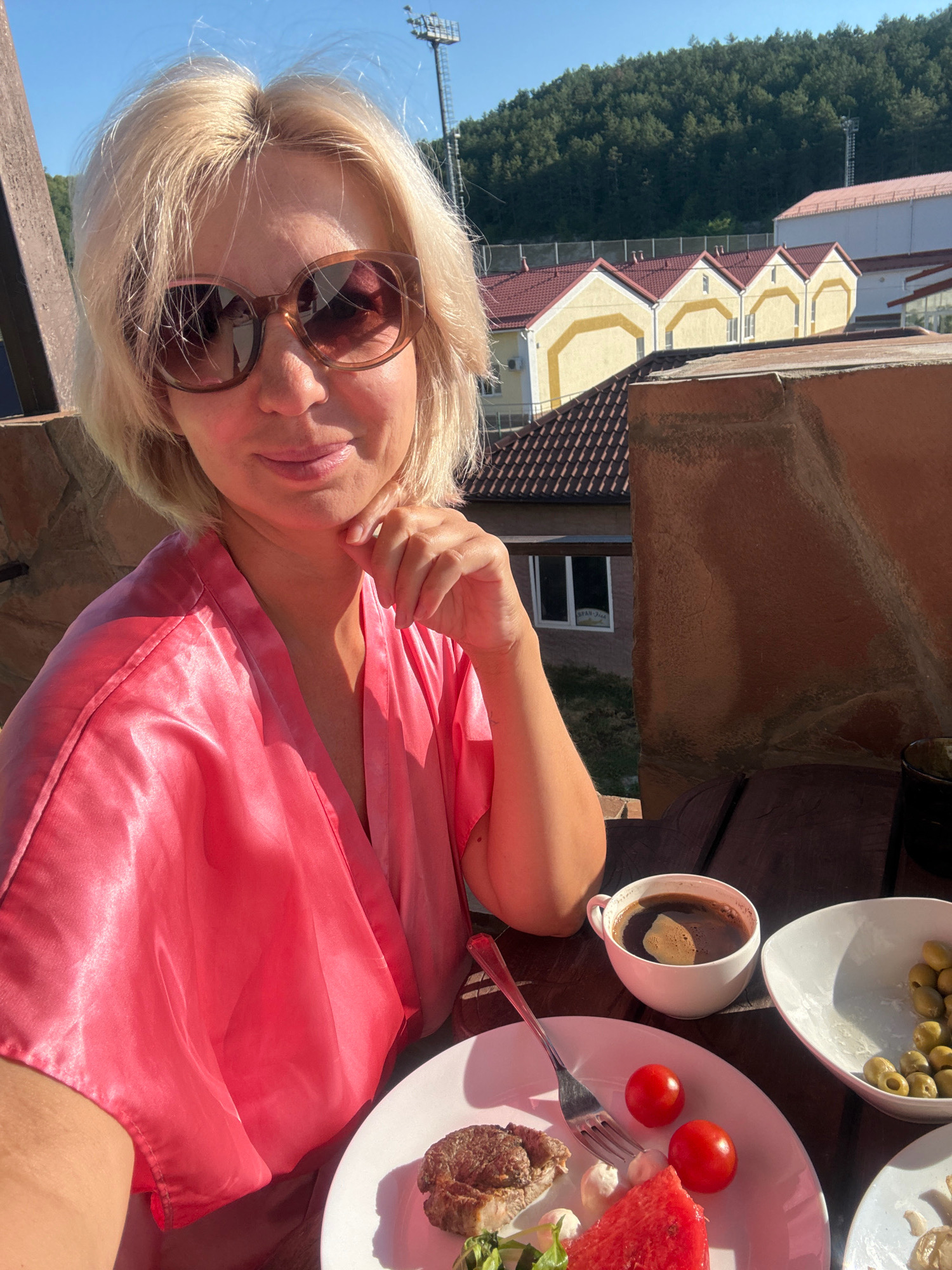 A woman with blonde hair and sunglasses is taking a selfie on a balcony overlooking a cityscape. She is wearing a pink satin robe. On the table in front of her is a plate with a steak, watermelon, cherry tomatoes, and cheese, a cup of coffee, and a bowl of olives. The background shows a row of houses with red roofs and a lush green forest. A tall tower with lights is visible in the distance. The sky is clear and blue. The image was taken in Abrau-Dyurso, Russia.