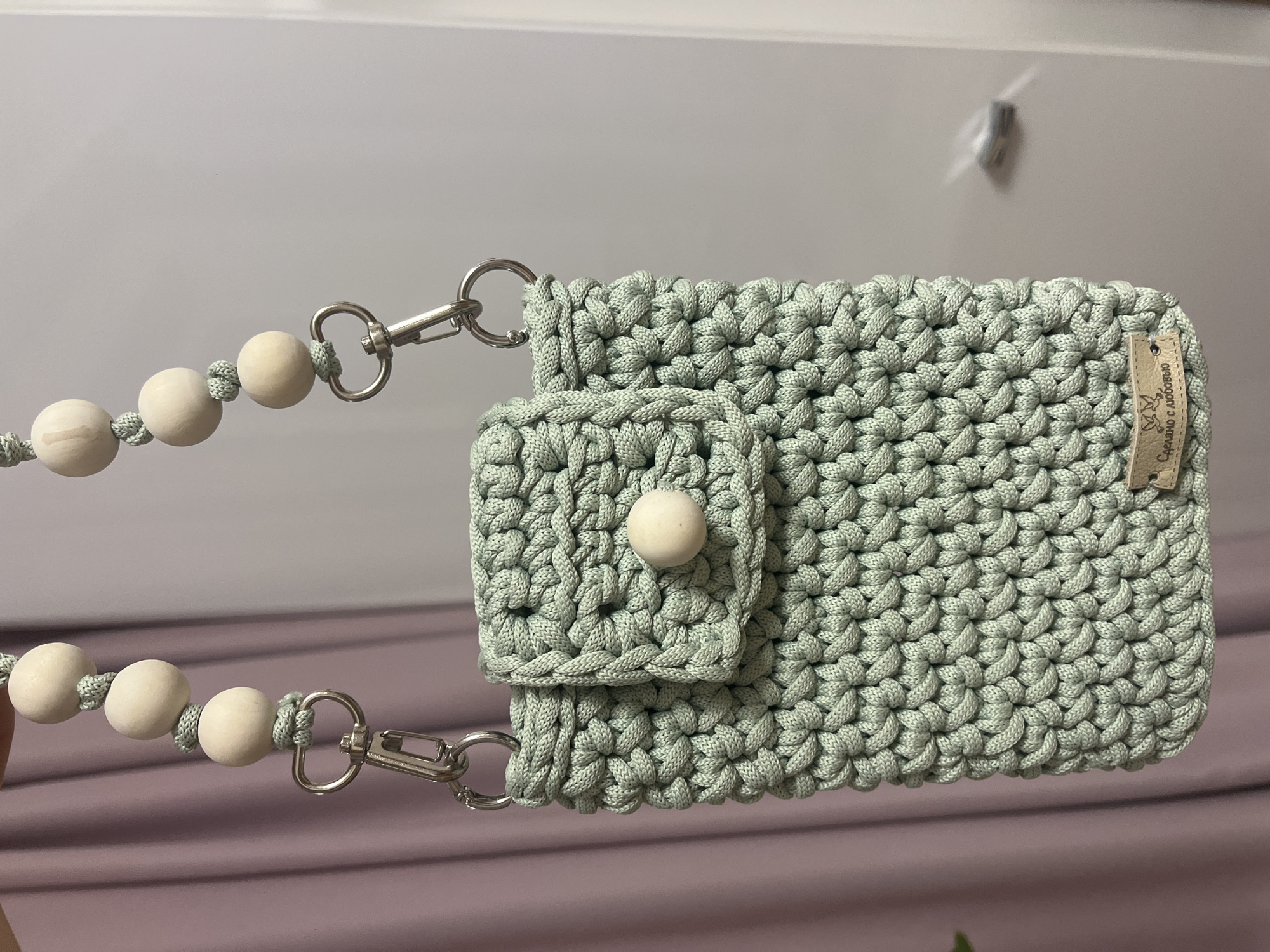 A close-up shot showcases a light green, crocheted wallet-purse featuring a flap with a wooden button closure. Attached to the wallet via silver-colored clasps is a string of wooden beads, evenly spaced. A small tag is stitched onto the side of the wallet. The background consists of blurred surfaces in white and mauve. The location is identified by the writing on the tag, written in Russian, that translates to "Made with love" and the city of Nizhny Novgorod, Russia.