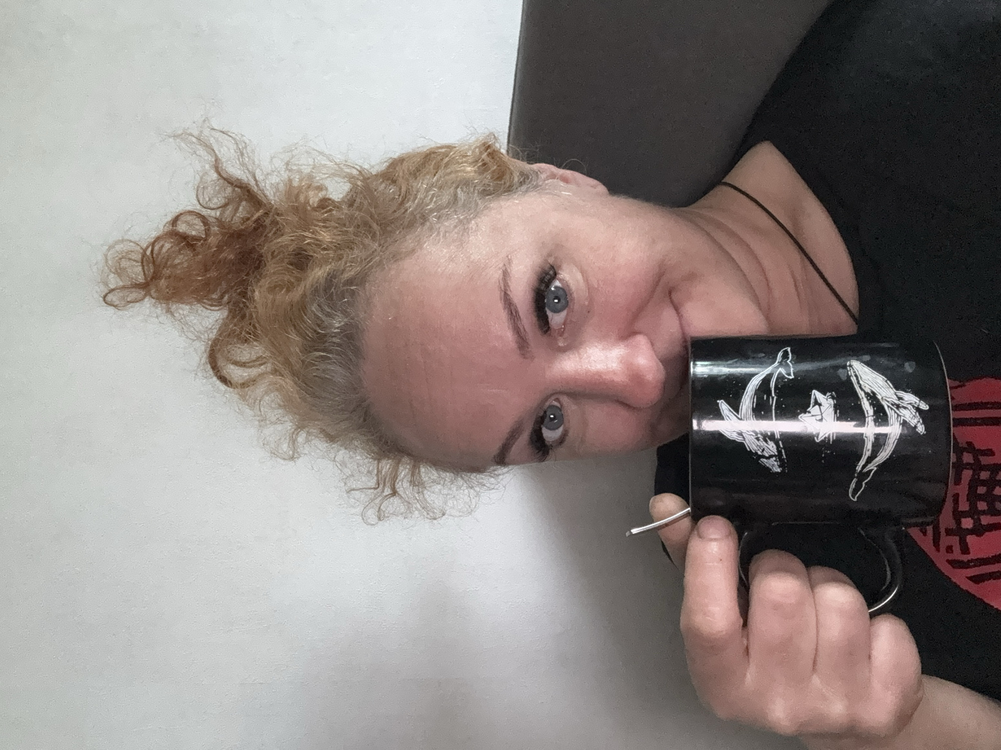A woman with light skin and light blue eyes is holding a black mug with a white design of two dolphins oriented head-to-head in front of her face. Her curly blonde hair is pulled back. She is wearing a black shirt and a thin black necklace. The background consists of a light gray wall. The image is rotated counterclockwise. There is no information provided that would indicate that the image was taken in Chaikovsky, Russia.