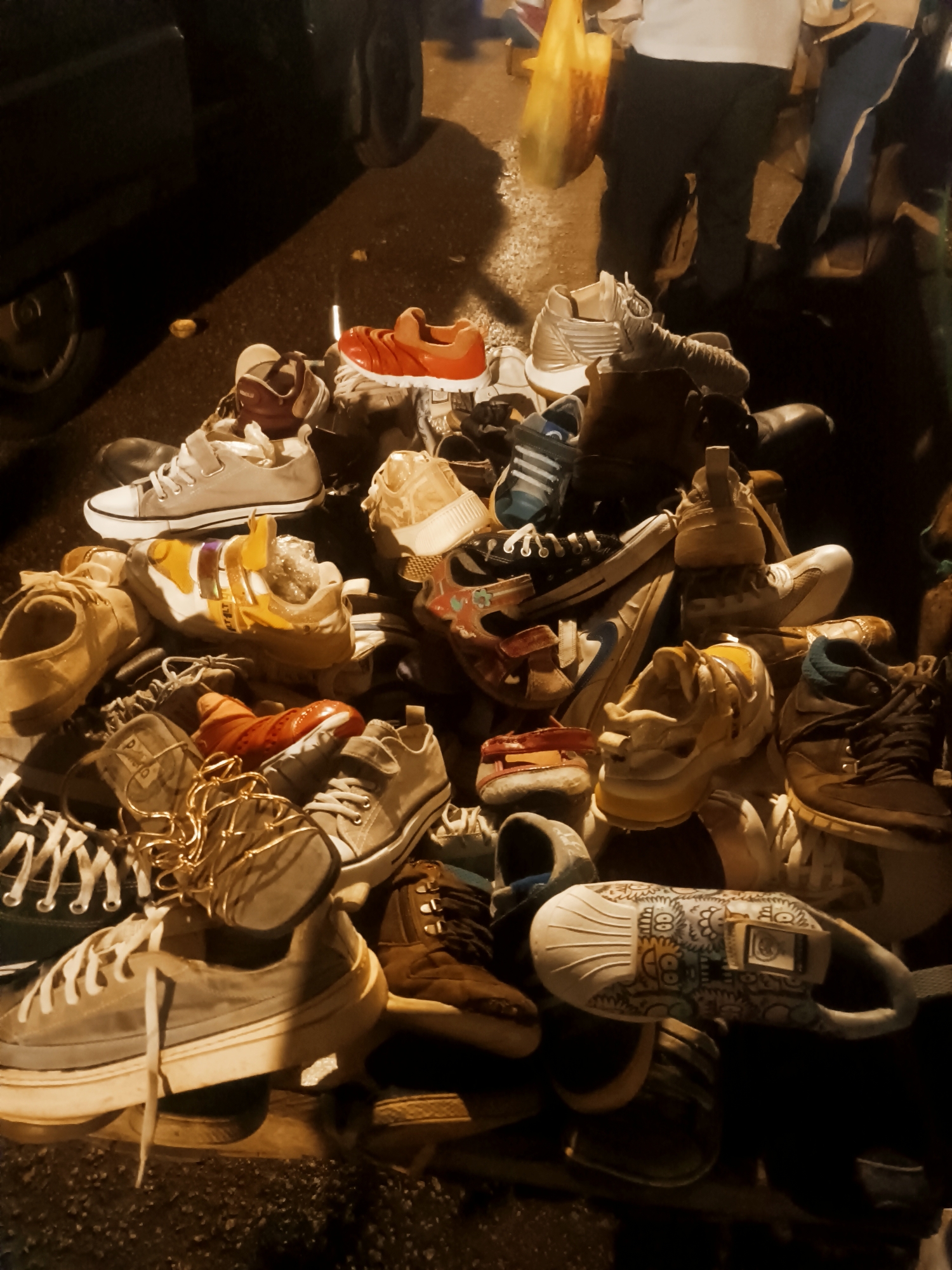 The image shows a pile of assorted shoes on the ground, seemingly for sale. Various types of footwear, including sneakers, sandals, and possibly boots, are present. The lighting suggests it might be evening or night. People are standing in the background, some holding bags. The location is likely an outdoor market or street vending spot in Bukuru, Nigeria.