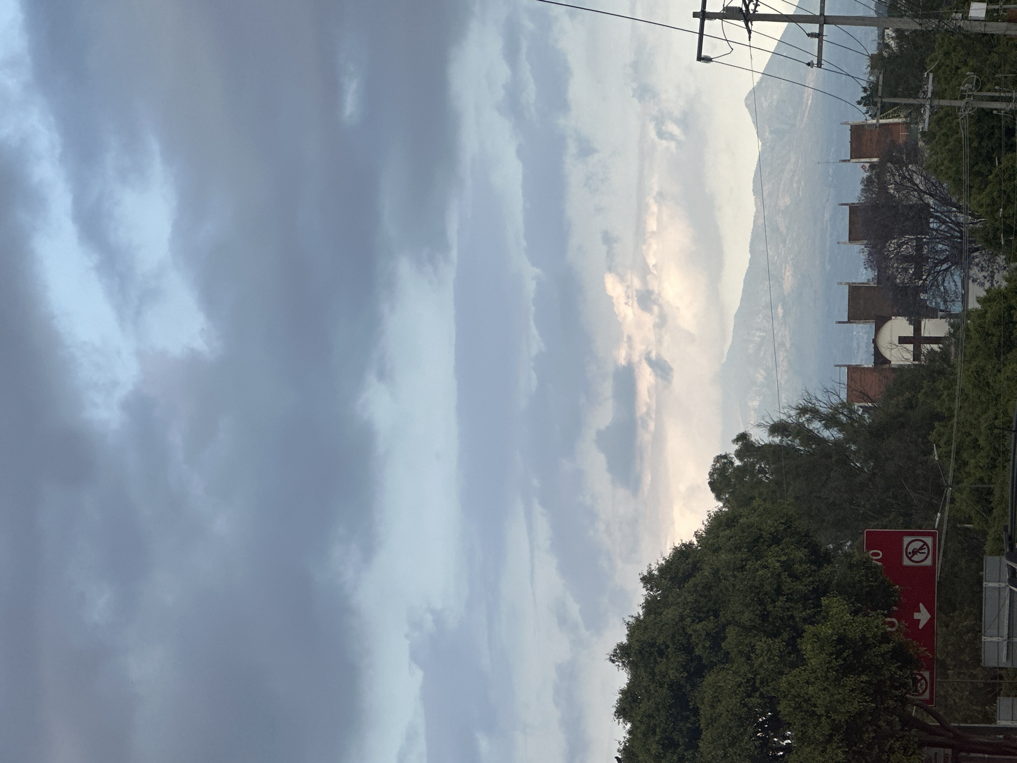 The image depicts a view of a landscape in the city of Unknown, Unknown. The sky is filled with dark, overcast clouds, with some lighter patches suggesting the sun is trying to break through. In the distance, there is a mountain range. A few buildings with orange-tiled roofs are visible, including one that appears to have a white cross on its facade. The foreground is dominated by lush green trees and foliage. A red sign is visible, featuring a pedestrian symbol with a line through it and an arrow pointing right, indicating a restriction or direction. Overhead, power lines and poles are present.