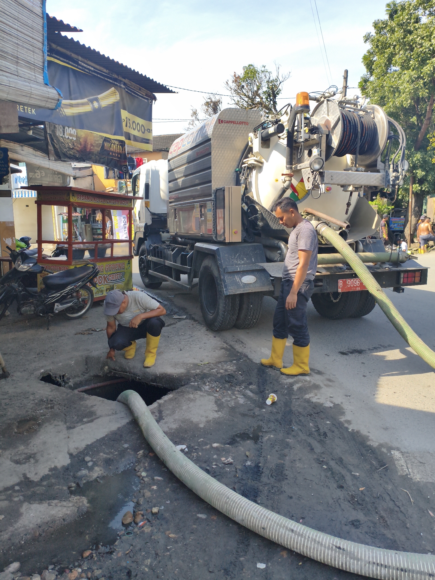 In the City of Tebing Tinggi, Indonesia, a street scene captures two men working near a large vacuum truck. The truck, a white Cappellotto model, is parked on the right side of the frame, its large tank and hose prominent. A green hose extends from the truck and lies across the asphalt, entering a manhole on the left.

One man, wearing a gray t-shirt, dark pants, and yellow rubber boots, is crouched by the manhole, appearing to inspect it. He is looking down into the opening. The second man, also in a gray t-shirt, dark pants, and yellow rubber boots, stands near the rear of the truck, looking down at the ground.

The environment is a street with a rough asphalt surface, marked by dirt, debris, and puddles of water. In the background, a small food stall with red and yellow awnings is visible, along with a parked motorcycle. Banners hang from buildings above the stall. The sky is bright and clear, indicating a sunny day. The license plate on the truck reads "B 9096 11-25".