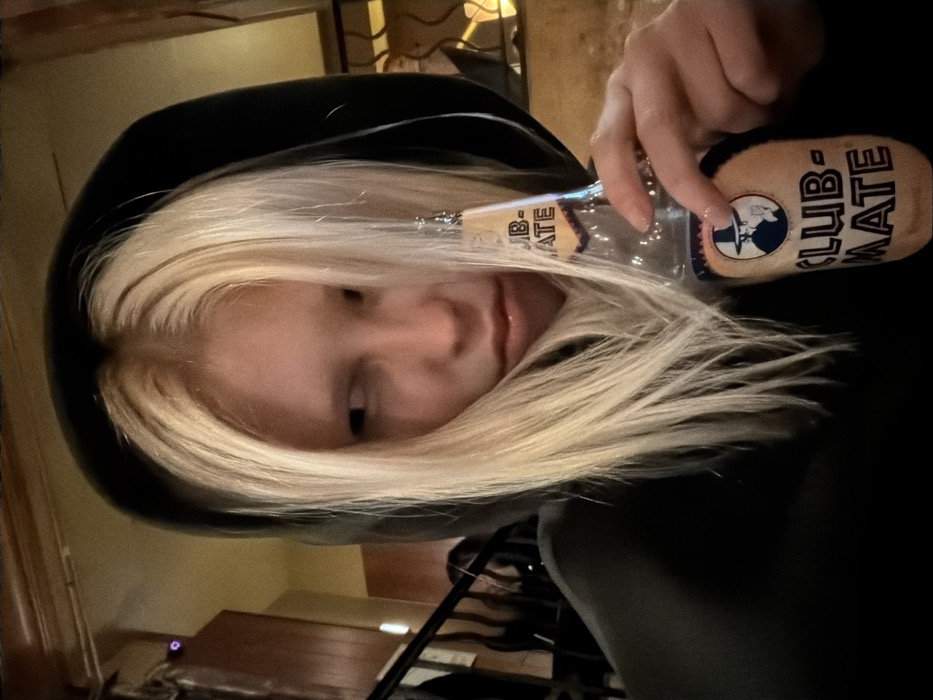 A person with blonde hair and a black hoodie holds a glass bottle of Club-Mate. The person's face is slightly tilted, with a focus on their expression. The bottle is prominently displayed, with the Club-Mate logo visible. The background appears to be indoors, possibly in a restaurant or bar. A table and other furniture are blurred in the background. The lighting is dim and creates a cozy atmosphere.