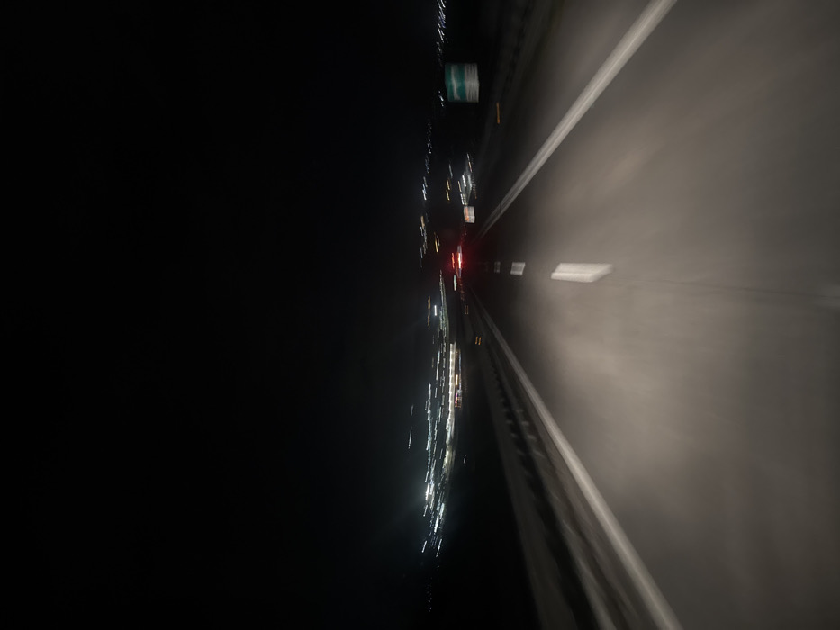 The image depicts a nocturnal scene from the perspective of a moving vehicle on a multi-lane roadway located within or near Campomarino, Italy. The asphalt road surface is visible, illuminated by an unseen light source, revealing distinct solid white lines marking the shoulders and dashed white lines indicating lane divisions. A metal guardrail runs along the left side of the road. The upper portion of the frame is dominated by a dark night sky. In the mid-ground, blurred streaks and points of artificial light on the horizon indicate a distant urban or populated area. Red vehicular taillights are discernible ahead on the roadway, along with other diffused white lights consistent with additional vehicle activity. A blurred, rectangular road sign with green content is partially visible on the right side of the road. The entire image exhibits significant motion blur, characteristic of a photograph captured from a vehicle traveling at speed during nighttime conditions. No individuals are visible.