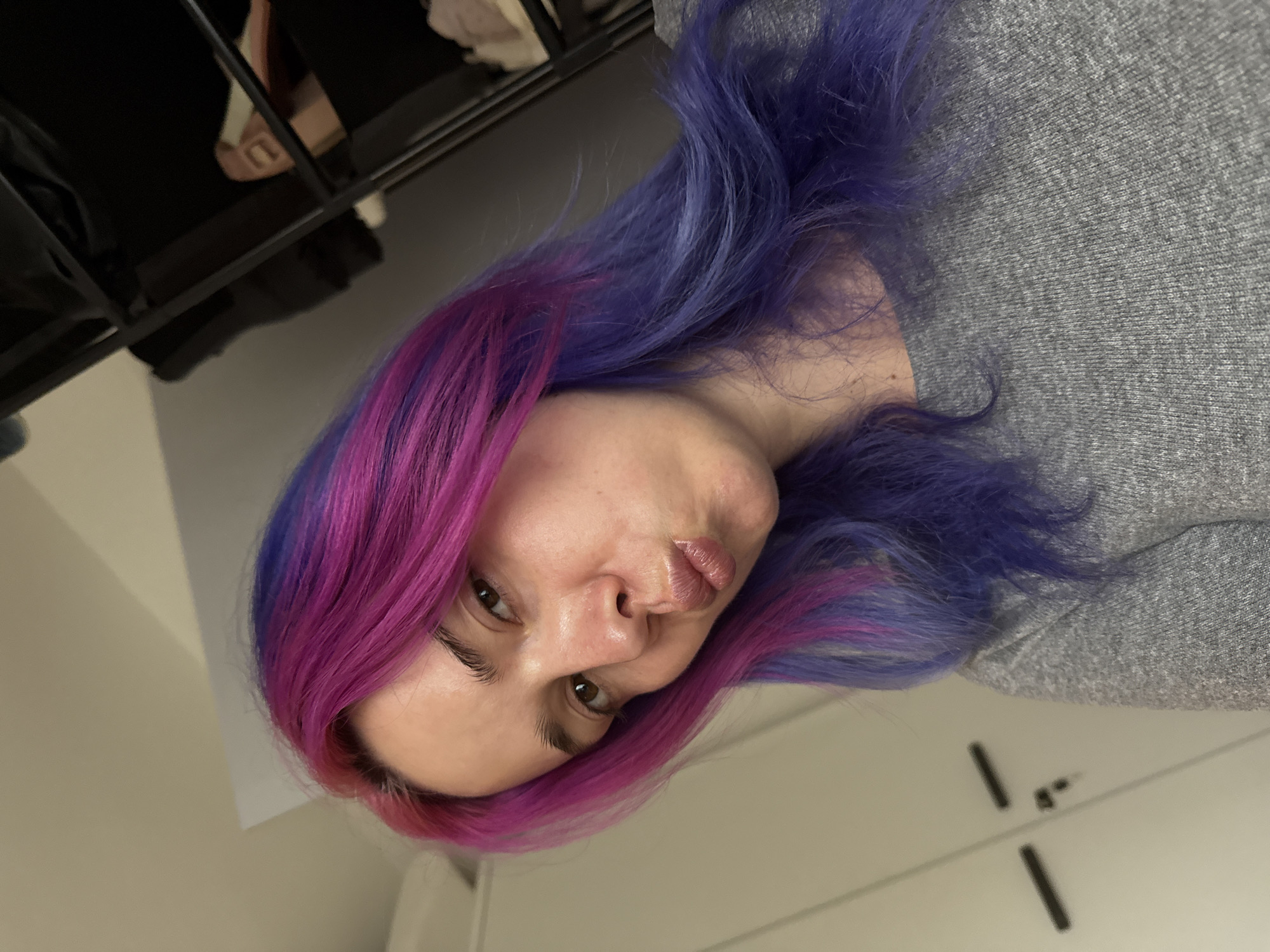 A woman with purple and pink hair is wearing a gray sweatshirt and making a kissing face. She appears to be indoors, in what seems to be a home or apartment. The location in the City of Zagreb, Croatia is undetermined.