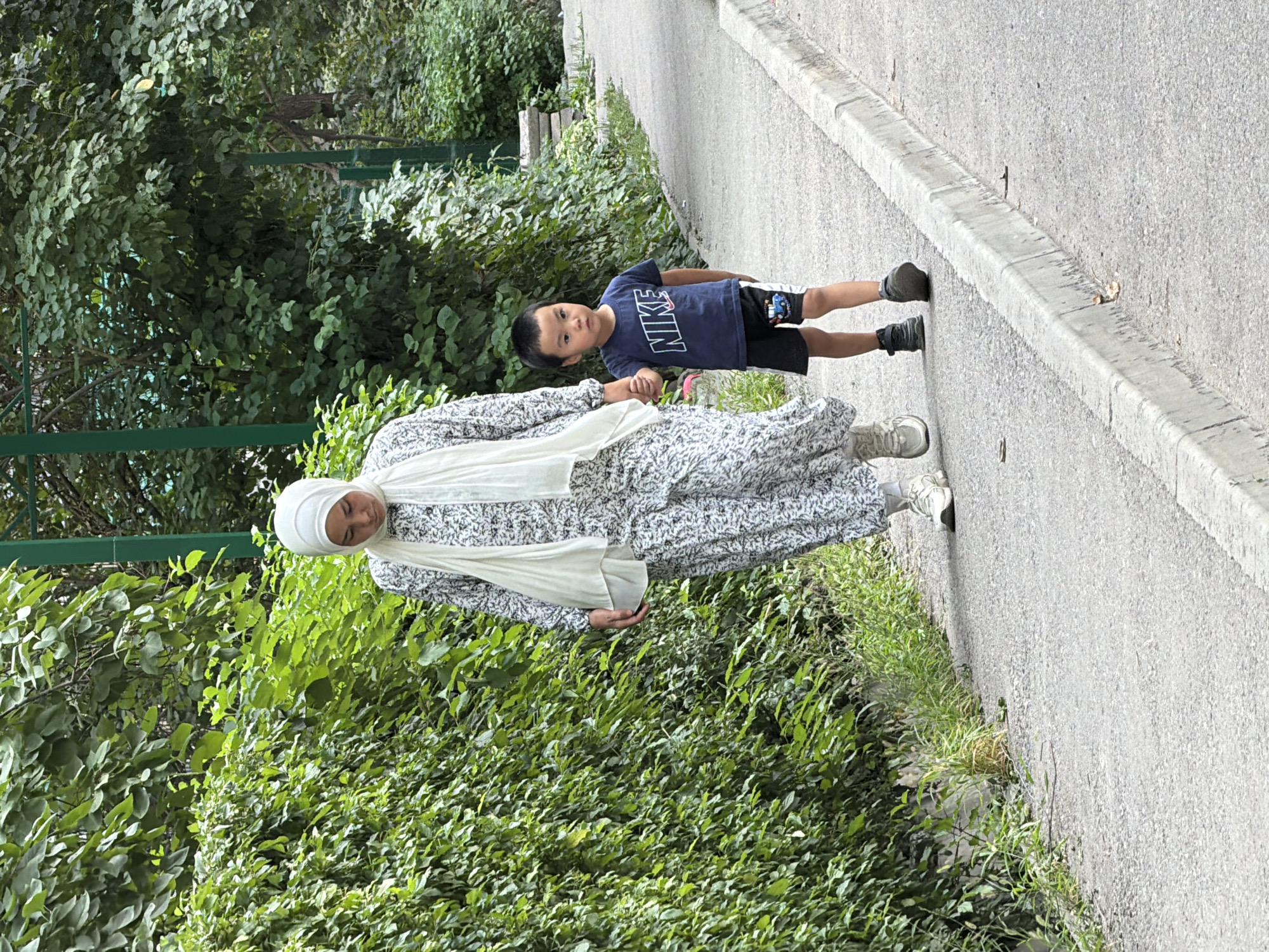 An adult woman wearing a white hijab and a patterned dress walks hand-in-hand with a young boy wearing a blue Nike t-shirt and black shorts. They are walking on a paved path lined with a concrete curb, bordered by dense green foliage on the left and a street or continuation of the path on the right. The woman is looking down, and the boy is looking forward with a neutral expression. The scene appears to be outdoors in a park or a tree-lined street.