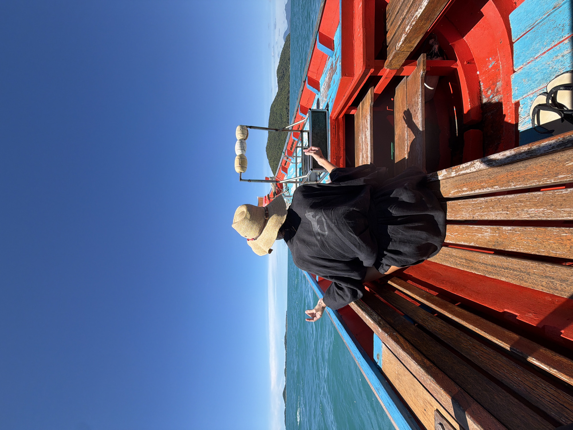 In Baan Thong Krut, Thailand, a person sits in a boat, facing away from the camera. They are wearing a straw hat and a black robe. Their hands are raised, palms facing upwards, in a meditative pose. The boat is red and blue, with wooden benches. The sea is a vibrant turquoise, and a clear blue sky stretches above. In the distance, lush green islands dot the horizon. The scene conveys a sense of peace and tranquility.