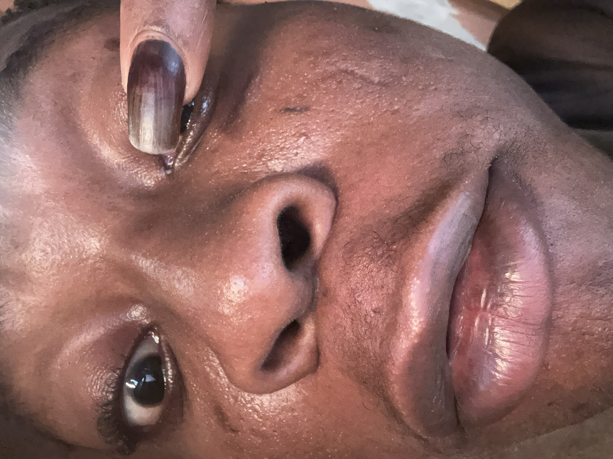 A close-up, tilted view of a dark-skinned individual's face is presented. The individual is lying down, with their face angled towards the camera. The left eye is open and visible, with a clear reflection in the pupil. A finger with a painted nail is near the left eye. The nose is prominent, and the lips are slightly parted. The skin appears to have some texture and moisture. No location is identifiable.