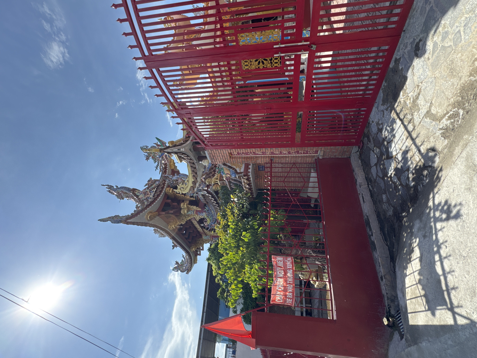 The image depicts the entrance to a Buddhist temple in Nha Trang, Vietnam, on a sunny day. A bright red gate dominates the foreground, with a smaller red gate to the left. Behind the gates, a vibrant, ornate temple structure with colorful dragon motifs is visible. A lush green tree stands in front of the temple. A red flag is planted to the left of the entrance, and a sign with Vietnamese text is attached to the fence. The ground in front of the gate is paved with stone, with shadows of the fence and foliage cast upon it. A drain grate is visible on the lower left side. The sky is a clear blue with a few wispy clouds and a bright sun shining through power lines. The overall scene suggests a well-maintained religious site in a bustling urban environment.