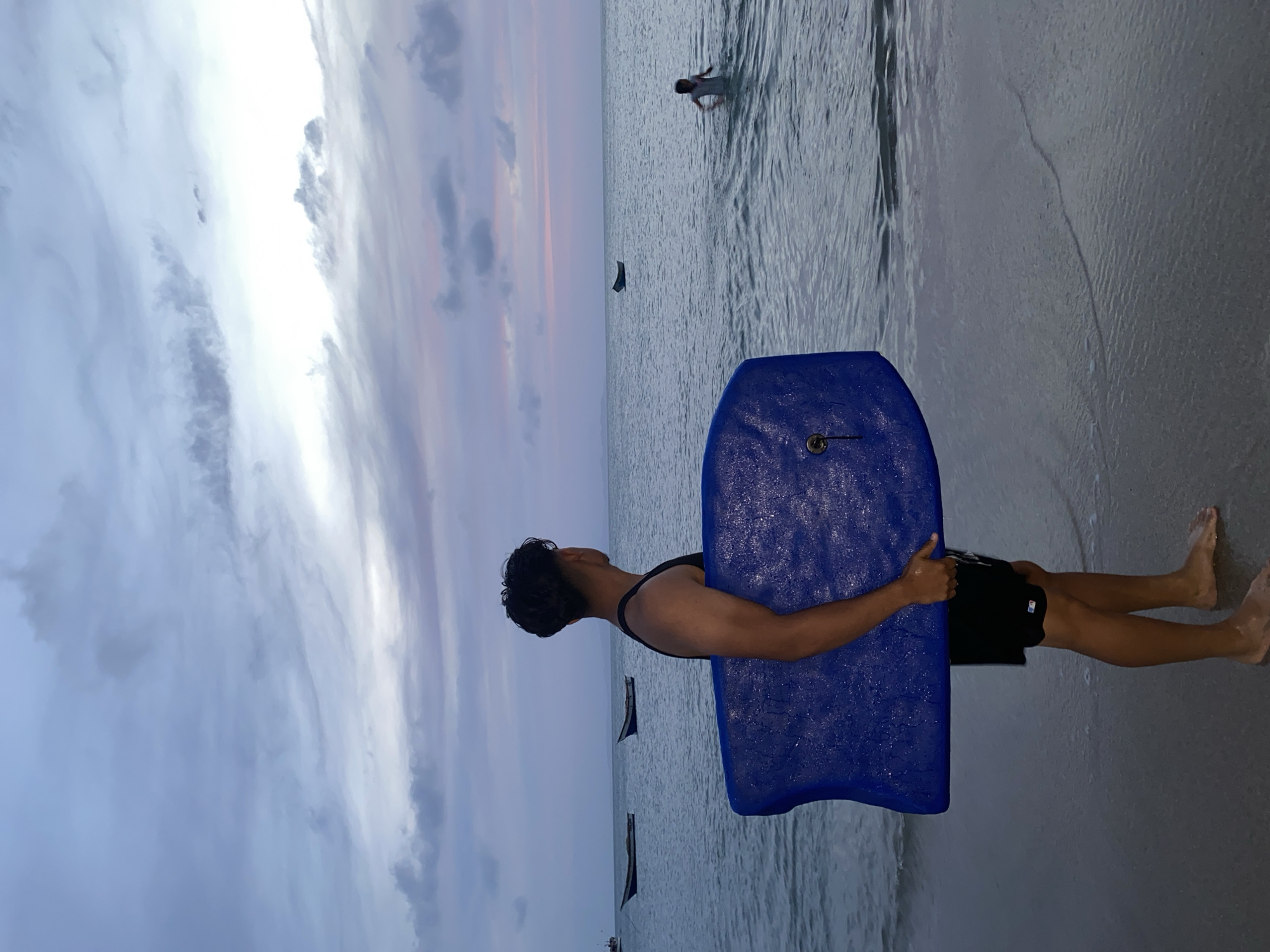 A person stands on a beach holding a blue boogie board. The individual is wearing a black swimsuit. The scene is likely in Juan Griego, Venezuela, based on the prompt. The beach has a wet, sandy surface and the ocean stretches out to the horizon, where the sky is filled with gray and light-colored clouds. In the distance, there are boats and a person is walking through the water.
