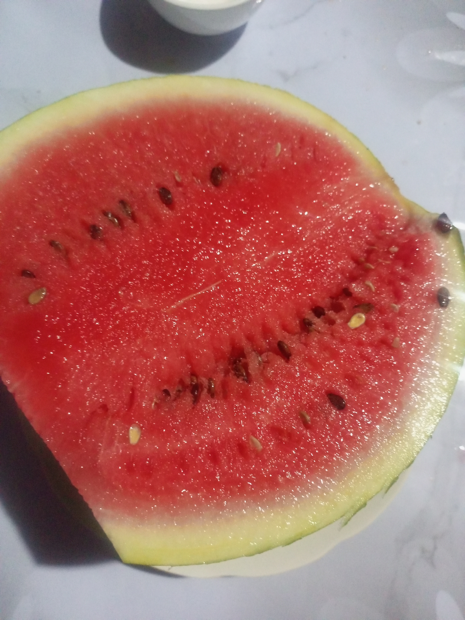 A close-up, overhead shot captures a slice of watermelon, its vibrant red flesh dotted with numerous dark seeds. The rind is a blend of light green and yellow, and a sliver of white bowl is visible in the upper portion of the image. The scene is set on a pale, textured surface, possibly a table.