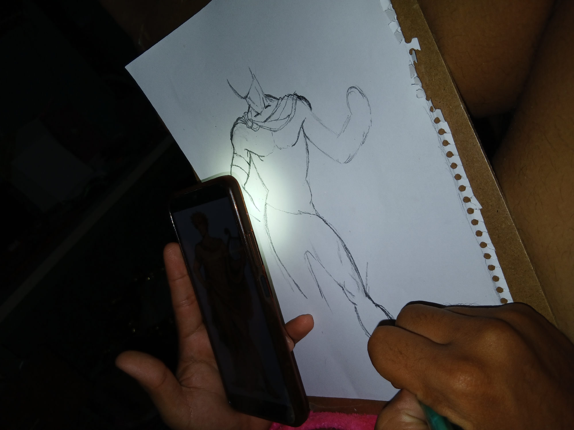 A person is drawing a figure on a piece of paper clipped to a clipboard. The person is holding a mobile phone in their left hand, using the phone's flashlight to illuminate the drawing. The figure appears to be a stylized, muscular character with a cape. The drawing is in progress. The location is unclear but possibly Altagracia, Venezuela.