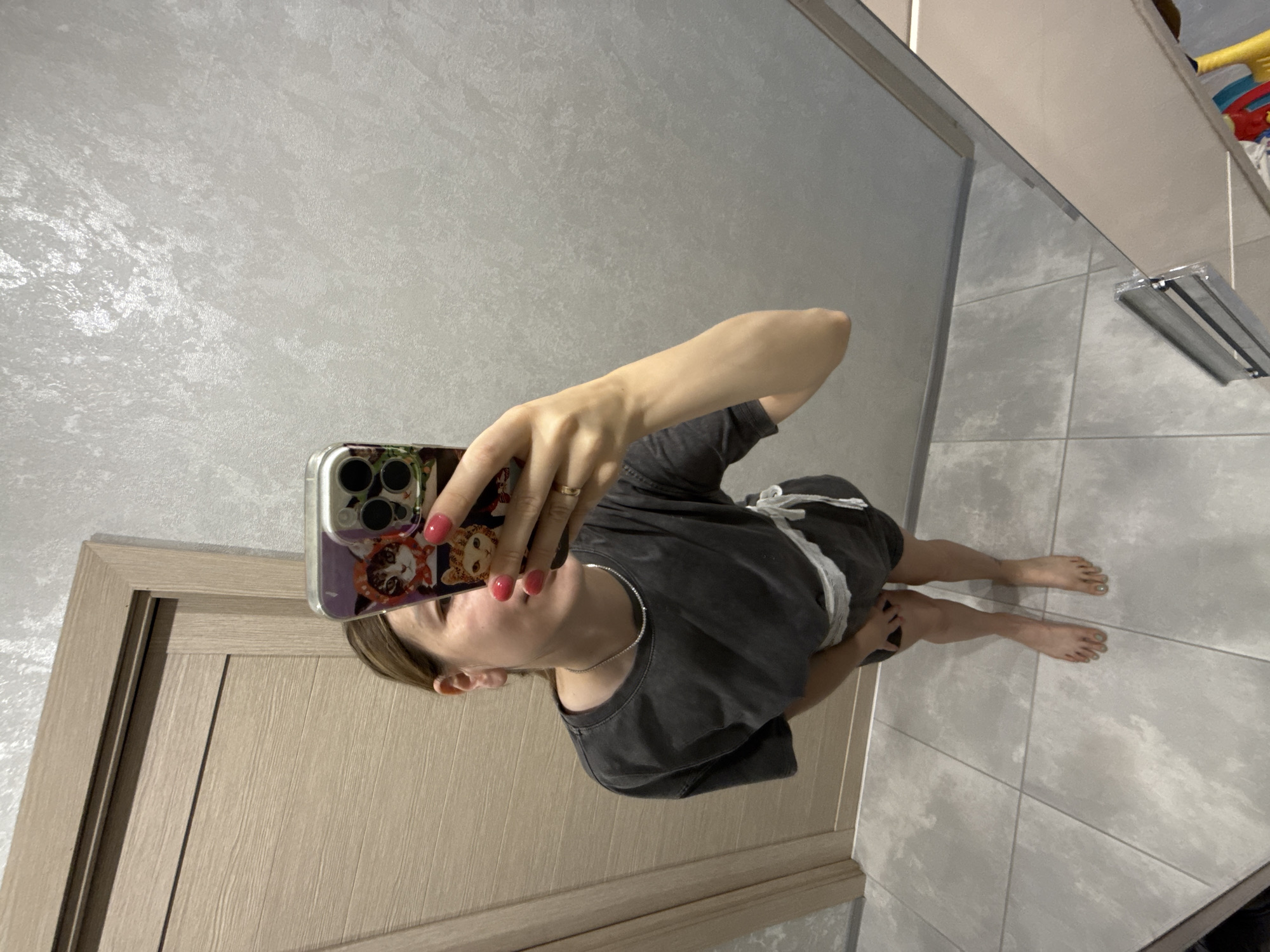 A woman is taking a mirror selfie in a room. She is wearing a dark gray t-shirt and shorts, with a white belt tied around her waist. Her nails are painted pink. She is holding a smartphone with a cat-themed case. The background includes a wooden door and a textured gray wall. The floor is tiled with large gray tiles. The lighting is bright, suggesting indoor lighting. The overall scene appears to be a domestic setting, possibly a bedroom or hallway. There is no indication of the location being Moscow, Russia.