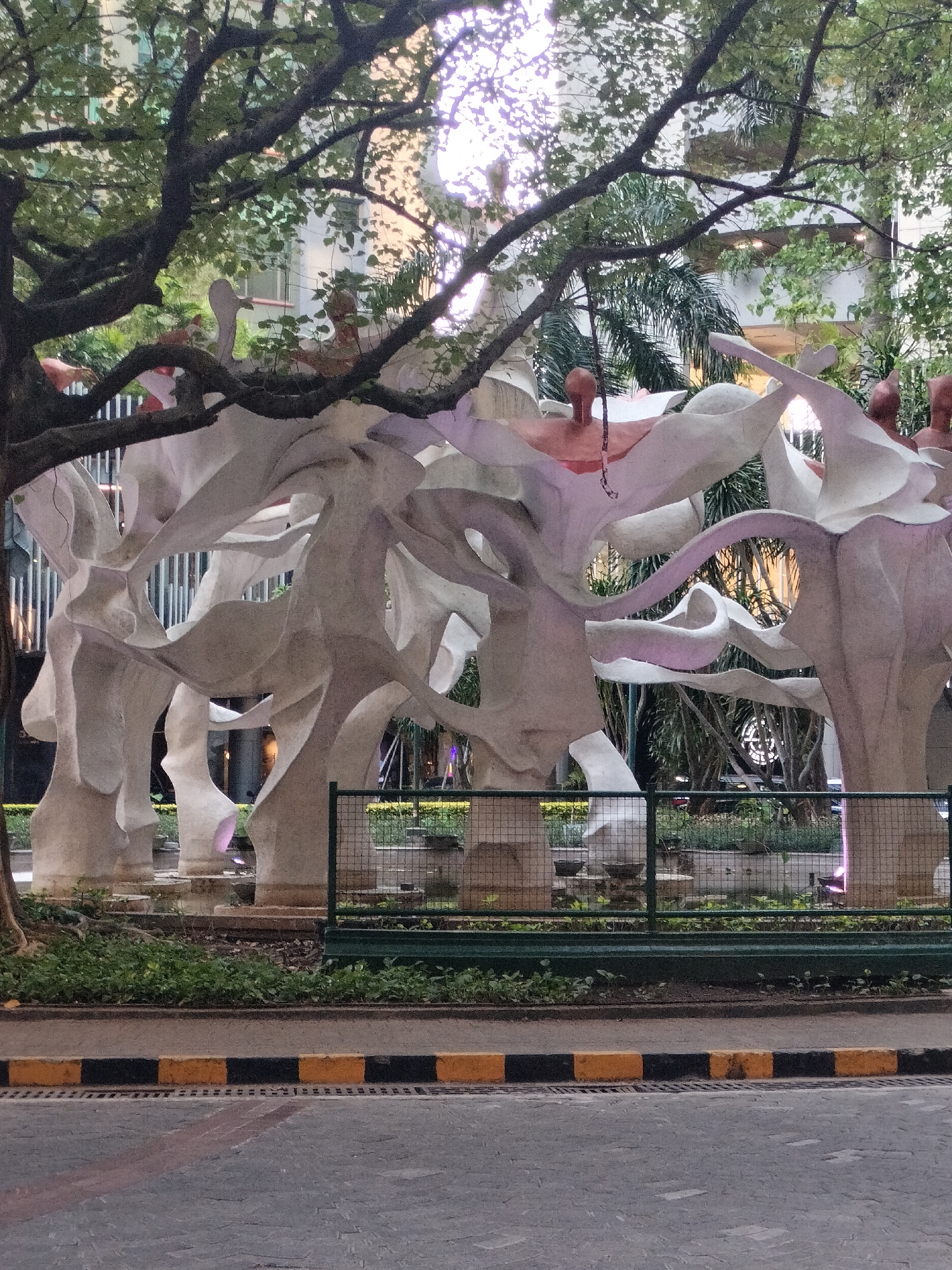 The image shows a large abstract sculpture with flowing, organic shapes, rendered in a pale, textured material. Several figures, appearing to be human torsos with heads, are integrated into the sculpture, some with visible arms. The sculpture is situated outdoors, with lush green trees and foliage partially obscuring the background. In the distance, a modern building with glass elements is visible. A dark green metal fence with a wire mesh is in the foreground, separating the sculpture from a paved area and a street with yellow and black striped curbs. This sculpture is located in front of the Ciputra World Surabaya complex in Surabaya, Indonesia.