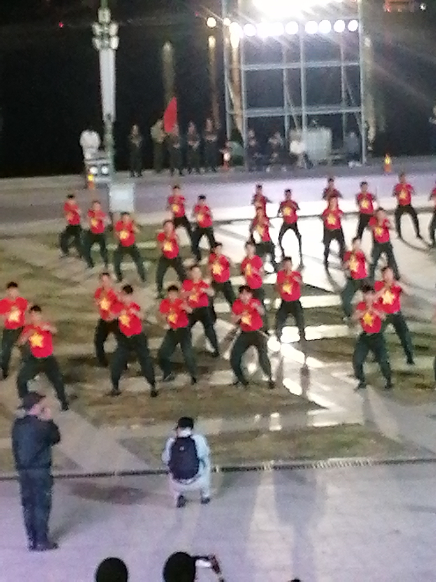 This media file depicts an outdoor event at dusk or night, likely in Da Lat, Vietnam. The central subjects are a large group of people, predominantly young men and women, dressed in matching red t-shirts with a yellow star emblem and dark pants. They appear to be performing a synchronized dance or routine on a paved surface, possibly a public square or stage area. The lighting is provided by stage lights, suggesting a performance or ceremony.
In the foreground, two individuals are more clearly visible. On the left, a person in dark attire, possibly a security guard or organizer, stands facing away from the camera. To the right, another person is crouched down, wearing a light-colored top and backpack, potentially observing or documenting the event.
Behind the performers, a large metal scaffolding structure is visible, illuminated by bright lights. Further in the background, indistinct figures and a red flag can be seen, hinting at a larger gathering or a patriotic theme. The overall atmosphere appears energetic and organized, given the synchronized movements of the performers. The time of day is clearly nighttime, indicated by the artificial lighting and the dark sky. There is no visible text in the image.