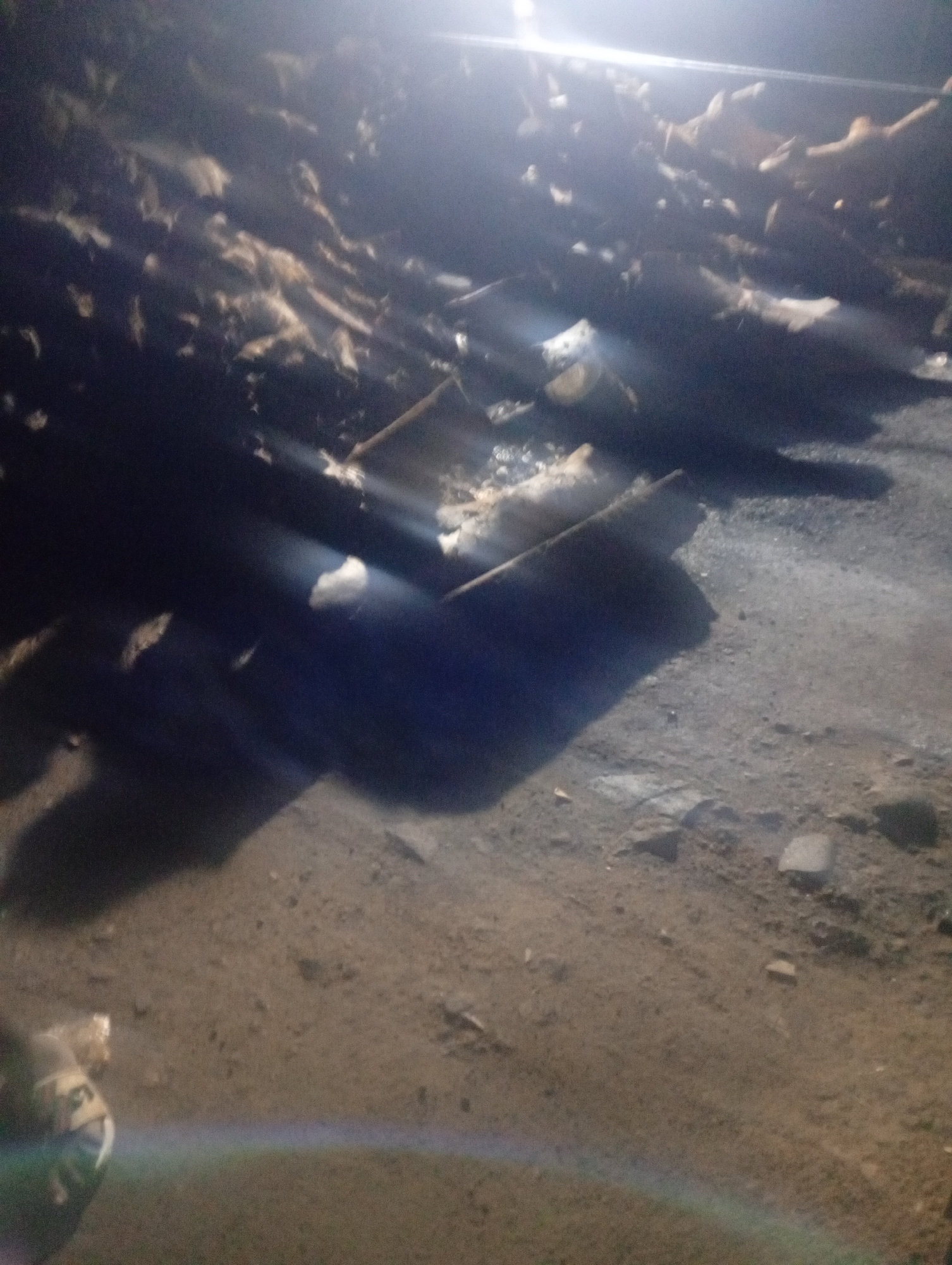 A nighttime scene in Bukuru, Nigeria, depicts a dimly lit outdoor area. The ground appears to be dirt or gravel, with scattered rocks. In the foreground, a person is partially visible on the left, wearing a dark garment. The middle ground and background are obscured by shadows and light flares from an overhead light source. There are various objects scattered across the ground, appearing as dark, elongated shapes, possibly debris or wood. Some lighter-colored material is also visible amongst the debris. The overall impression is one of disarray in a poorly lit outdoor environment.