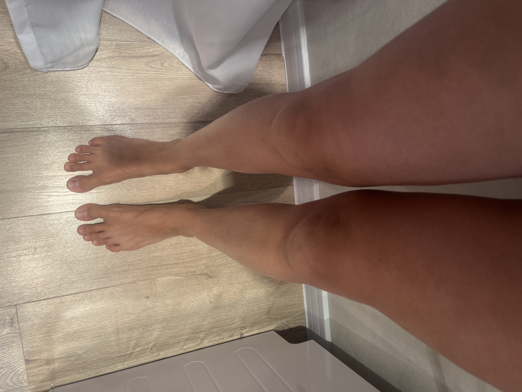 A low-angle, full-frame shot shows a pair of bare legs and feet standing on a light-colored wooden floor. The legs are tanned, and the feet are positioned slightly apart. To the left of the image, a white garment, possibly a dress or robe, is partially visible, draped on the floor. The background consists of a white baseboard and a textured wall. The overall scene suggests an indoor environment, possibly a residential setting in Rybnitsa, Moldova.