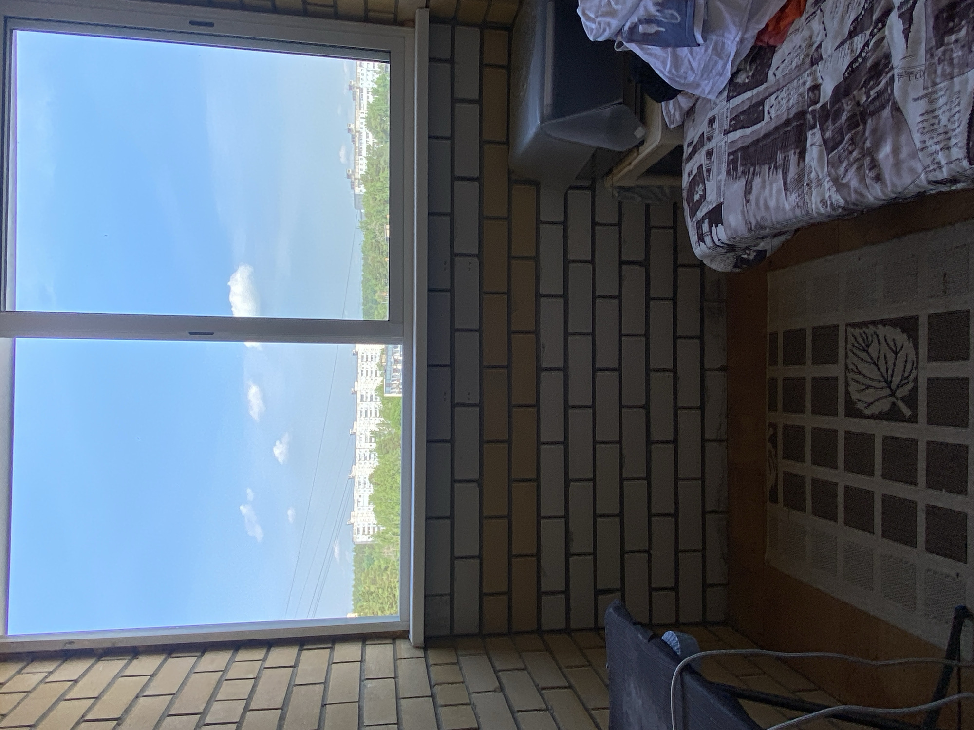 This media file shows a view from a balcony or a room with a large window. Through the window, a bright blue sky with scattered white clouds is visible, with a distant cityscape and a line of green trees below. The buildings in the distance are white and appear to be multi-story.
Inside the room, the wall is made of light-colored bricks, a common material for balconies in some regions. To the right of the window, a bed is visible, partially covered with a patterned duvet featuring a black and white design. Some clothing, including white and red fabric, is draped over the bed or a nearby shelf. A dark-colored object, possibly a piece of furniture or a storage container, is positioned in the lower right corner of the frame. The flooring appears to be tiled.
The lighting suggests it is daytime, likely late morning or early afternoon, given the bright sky. The weather appears to be clear and sunny. There is no visible text in the image. The overall scene conveys a sense of an indoor space with a view of an urban environment on a pleasant day.