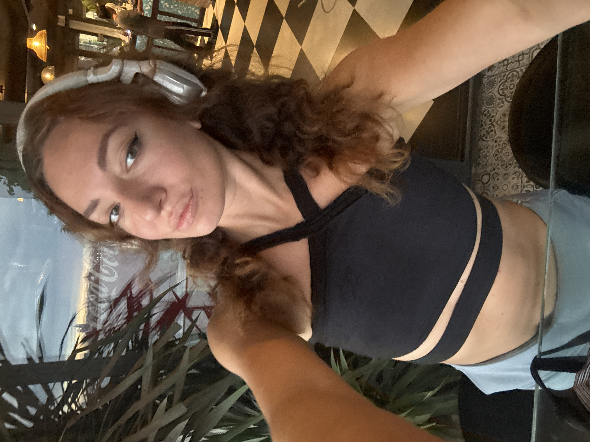 In an indoor setting, a woman with long, curly brown hair and headphones is captured in a selfie. She is wearing a black crop top with a halter neckline and light blue shorts. Her facial expression is neutral, with dark eye makeup. Behind her, a large window reflects an outdoor scene with plants and a Coca-Cola logo. The interior features a black and white checkered floor and a person in the background. The scene appears to be a cafe or bar. There is no indication that the image was taken in Hanoi, Vietnam.