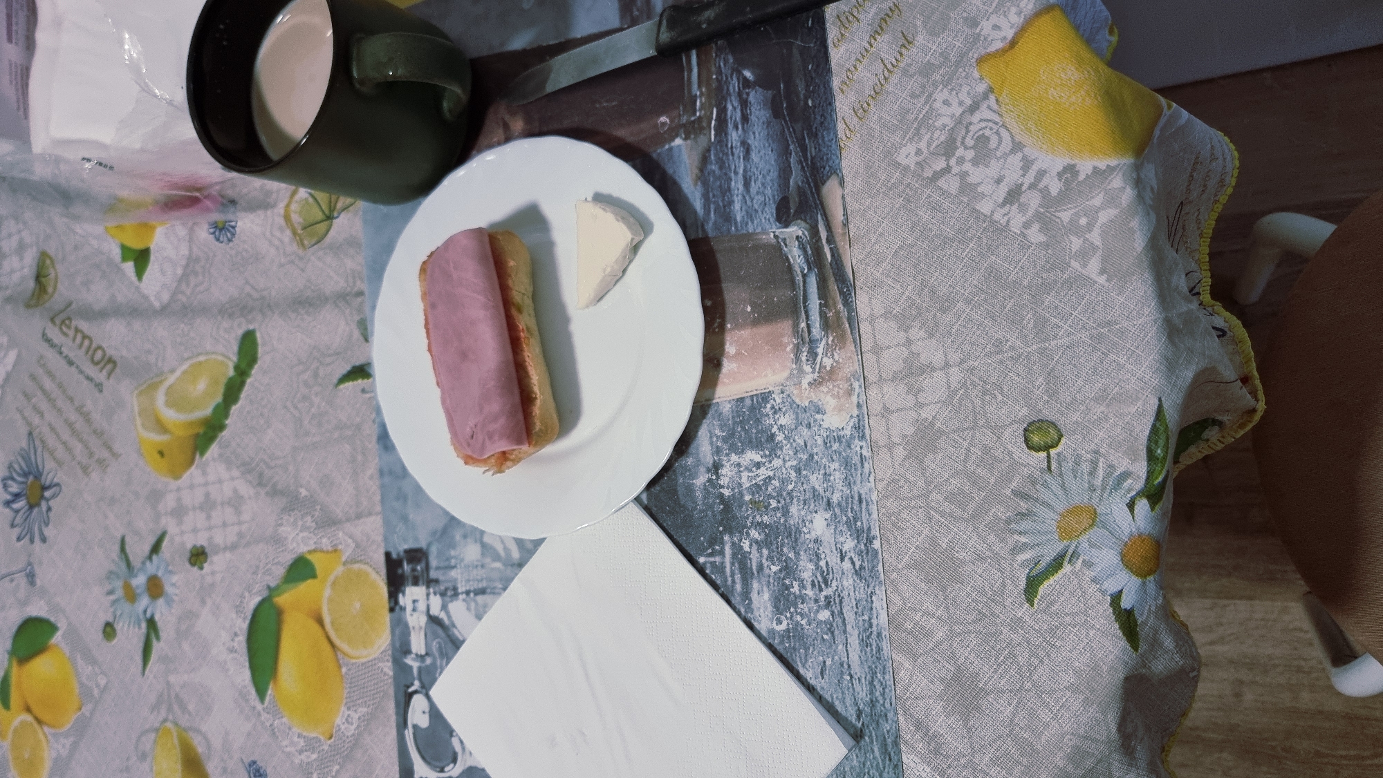 A close-up, overhead shot shows a breakfast setting on a table covered with a tablecloth featuring a lemon and daisy pattern. A white plate holds a sandwich topped with pink ham and a small triangle of white cheese. To the right of the plate is a dark green mug filled with a light brown beverage. A white paper napkin is folded neatly to the left of the plate. A knife with a dark handle lies to the right of the mug. The background of the tablecloth displays a faded image of what appears to be a glass. The scene is likely indoors, possibly in a home or cafe, in Sant Joan Les Fonts, Spain.