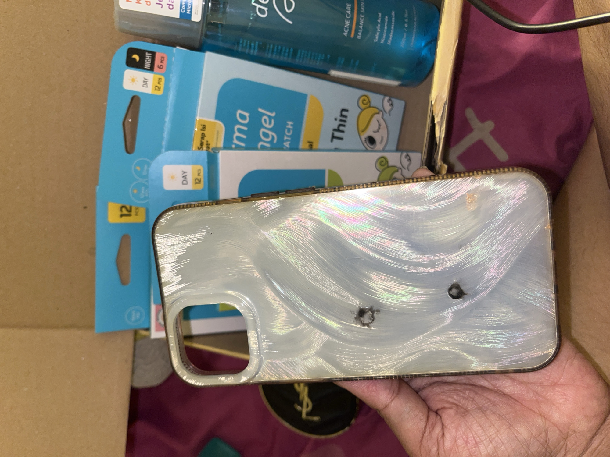A person is holding a silver iPhone case with two visible dark spots or holes on its surface. The case is above a cosmetic compact with the YSL logo. Around these items are cardboard boxes, a blue bottle, and blue and white packaging for acne patches. The patches are labeled "Derma Angel Thin" with "DAY 12 PCS" and "NIGHT 6 PCS" visible. The bottle appears to be a skincare product, featuring a blue liquid, and "ACNE CARE BALANCE SKIN" on the label. The background shows a glimpse of a purple fabric and the person's arm. Based on the product labels, it is likely located somewhere in Central Jakarta, Indonesia.