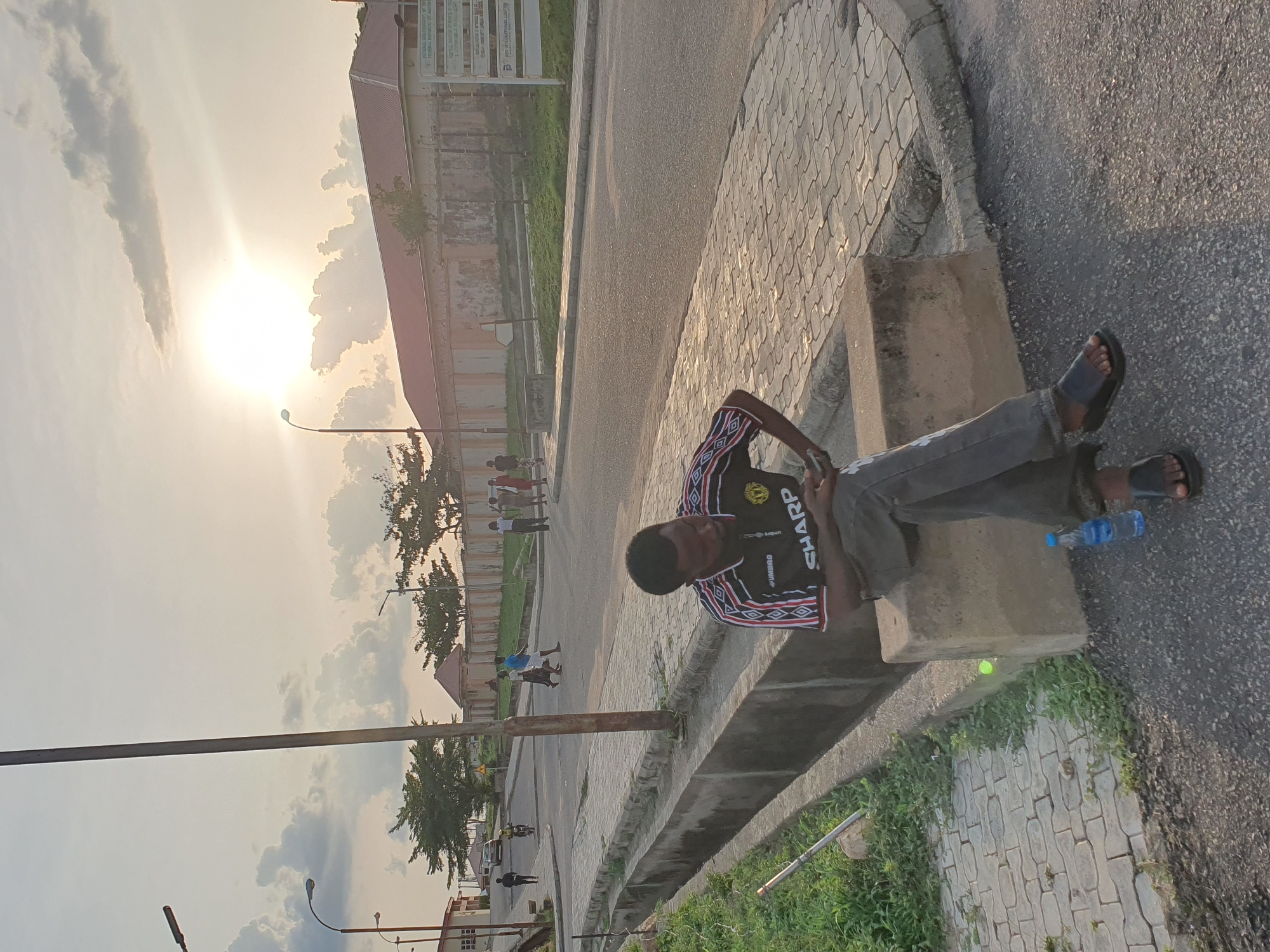 A person is sitting on a concrete structure along a roadside. The person is wearing dark-colored clothes, including a black jersey with the word "SHARP" on it and sandals. A water bottle is on the ground next to the person. The road is paved, and there are brick pavers on one side. In the background, there are buildings and trees, illuminated by a bright sun and partially obscured by clouds. In the distance, other individuals are walking. The location is likely Lokoja, Nigeria, given the context.