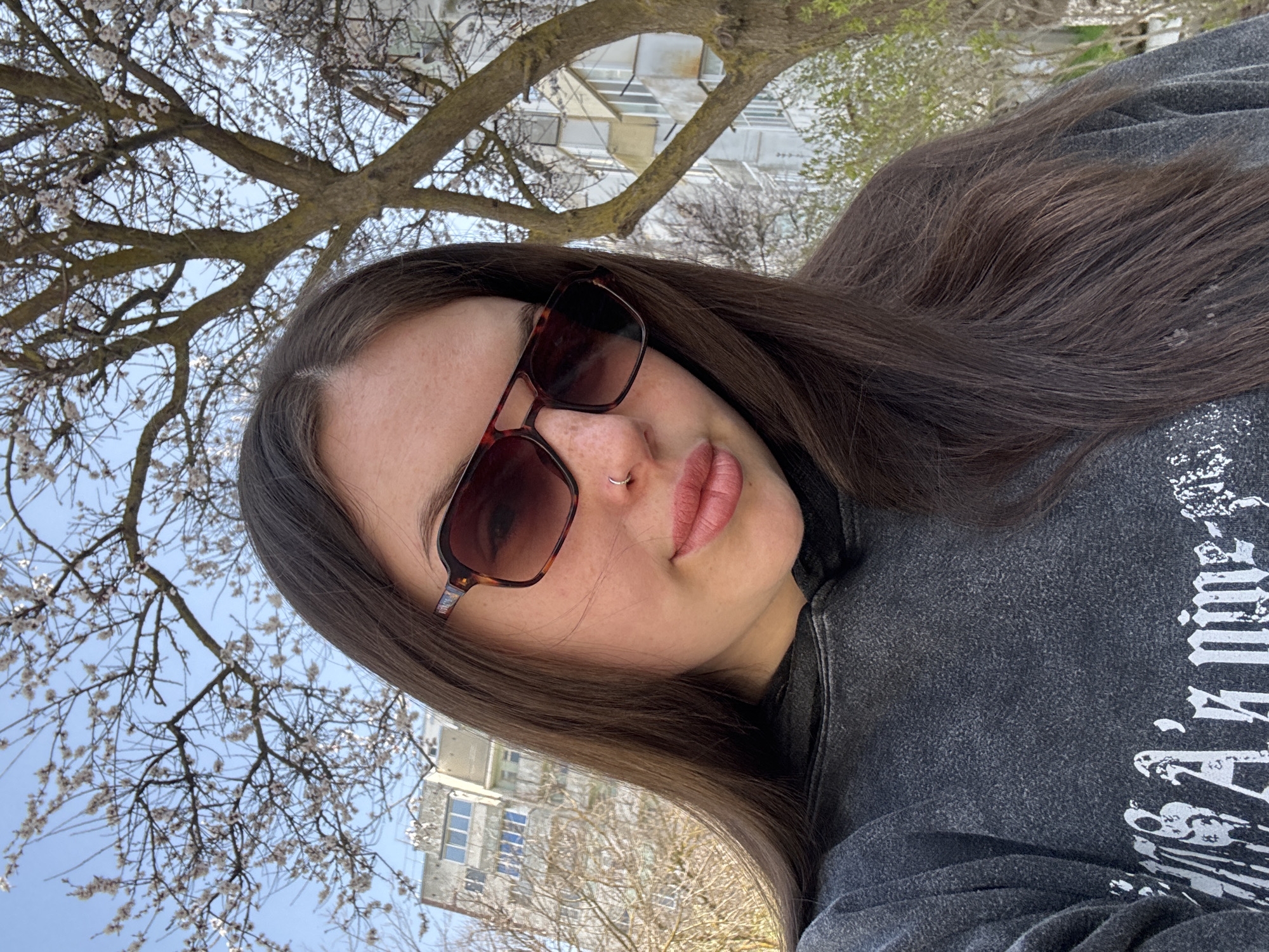 A person with long, dark hair and sunglasses is taking a selfie outdoors. They are wearing a gray hoodie with a white design on it. The person has a nose ring. Behind the person is a tree with bare branches and small white flowers. In the background, there is a light-colored multi-story building. Based on the description, the location is Chornomorskoe, Ukraine.