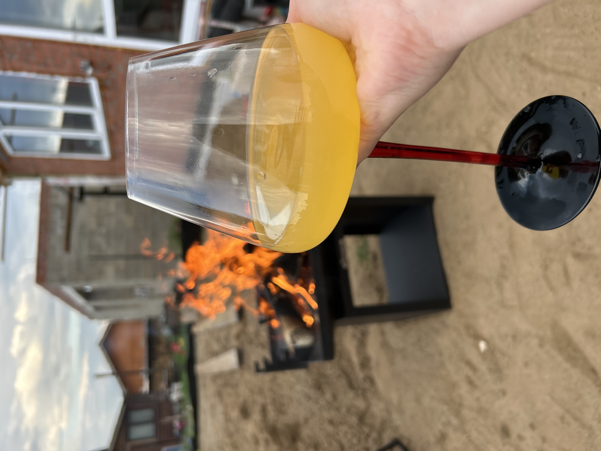 In the city of Chaikovsky, Russia, an outdoor scene unfolds with a close-up on a hand holding a wine glass filled with yellow liquid, possibly juice or a cocktail. The glass has a red stem and a black base. In the background, a black grill with a lively fire burning is visible. Beyond the grill, the landscape features partially constructed brick buildings, a wooden house, and sandy ground. The sky above is cloudy, suggesting an overcast day.