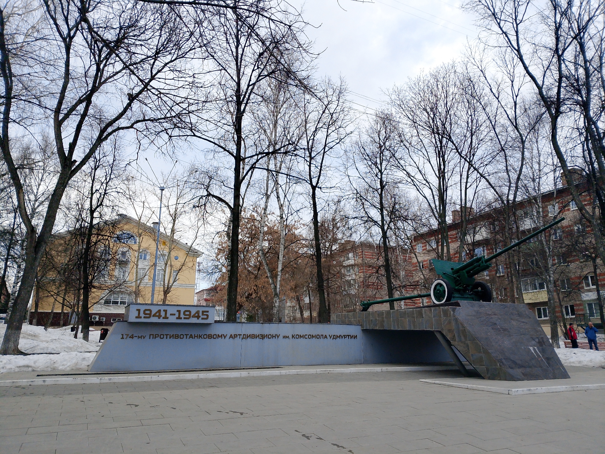 The image depicts a memorial complex in Izhevsk, Russia, commemorating the 174th Anti-Tank Artillery Division of the Komsomol of Udmurtia (1941-1945). A green anti-tank gun is mounted on a raised platform made of dark stone or concrete, which slopes down on either side. The platform is supported by a lower structure, creating a tunnel-like space underneath. The memorial's inscription is in Russian and includes the dates "1941-1945". Bare trees surround the memorial, indicating a winter or early spring setting. A yellow building is partially visible behind the memorial to the left and a reddish-brick building is visible behind it to the right. Patches of snow are visible on the ground. Two people are standing in the background to the right, near a red bench, and appear to be observing the monument. The paved area in front of the memorial suggests it is located in a public square or park. The sky is overcast.