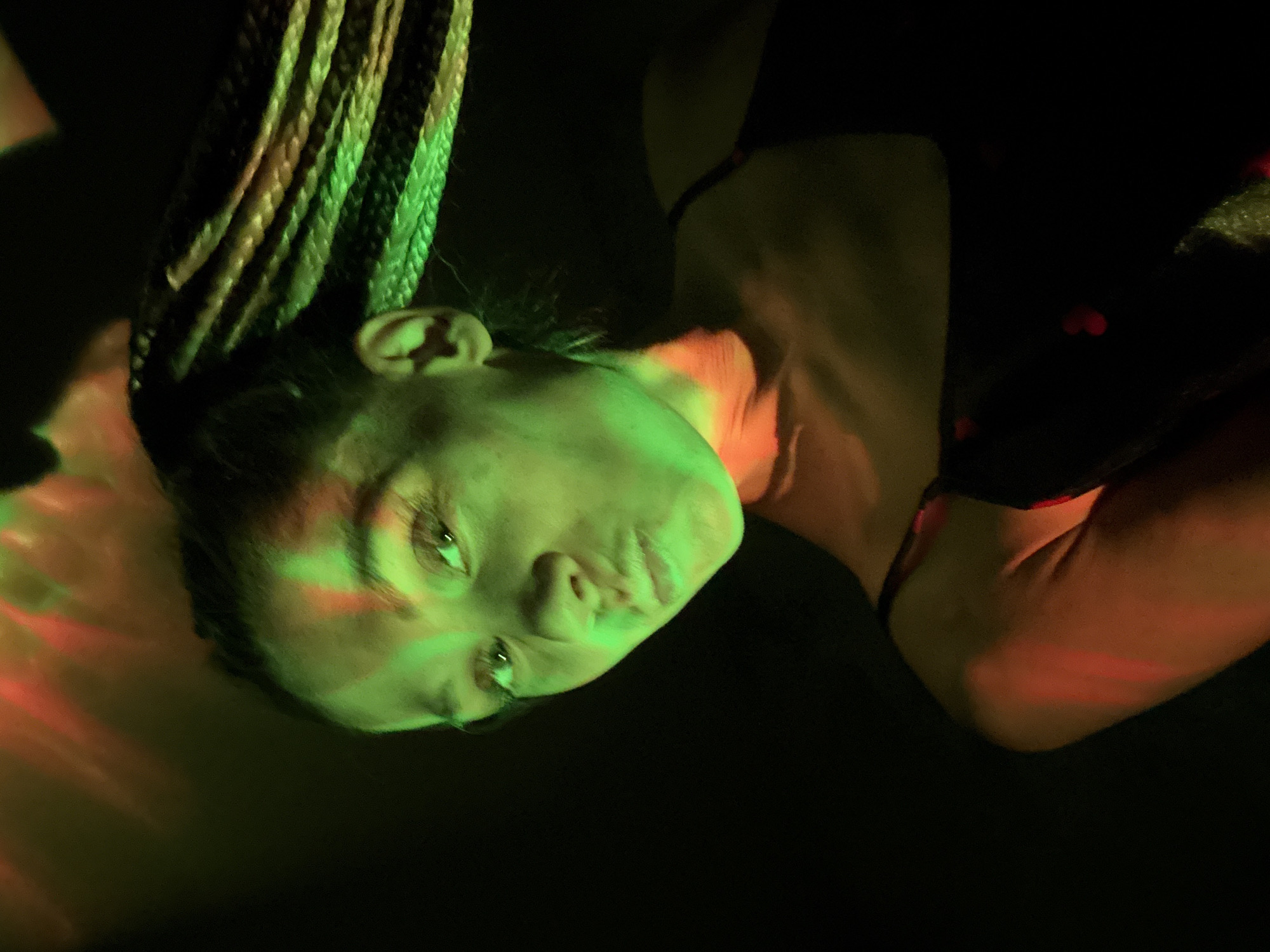 A woman with braided hair is depicted in the dark, under a green light illuminating her face and part of her braids. Red light is cast on her neck and shoulder. She has a piercing in her nose and is wearing a dark top with red hearts. The background is dark and indistinct. The location and date are unknown.