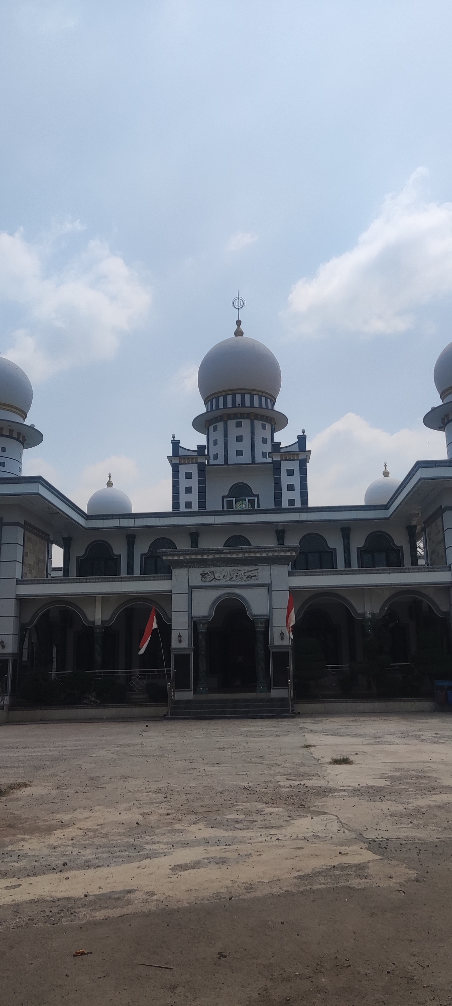 The image shows the exterior of a mosque in Panongan Lor, Indonesia. The mosque has a white facade with a prominent central dome, flanked by smaller domes. The architecture features arched windows and colonnades. Two Indonesian flags are visible, one on each side of the main entrance. The ground in front of the mosque is a dirt and concrete area. The sky above is partly cloudy. There are no people or animals visible in the image.