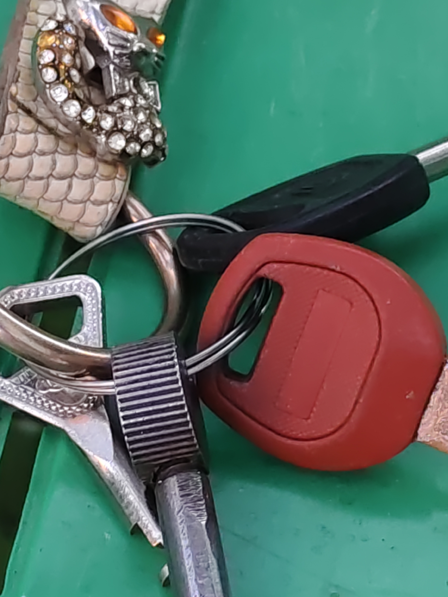 This image shows a close-up of a keyring with several keys and decorative charms. The keyring is on a green surface, likely a table or a bench. One of the charms is a silver skull with amber eyes and numerous small, clear rhinestones. Another charm appears to be a silver buckle with intricate detailing. There are two keys visible: one with a black plastic head and a silver shaft, and another with a red plastic head and a silver shaft. The green surface appears to be outdoors, possibly in a park or public area in Rostov-on-Don, Russia, given the context provided. There are no people or actions depicted.