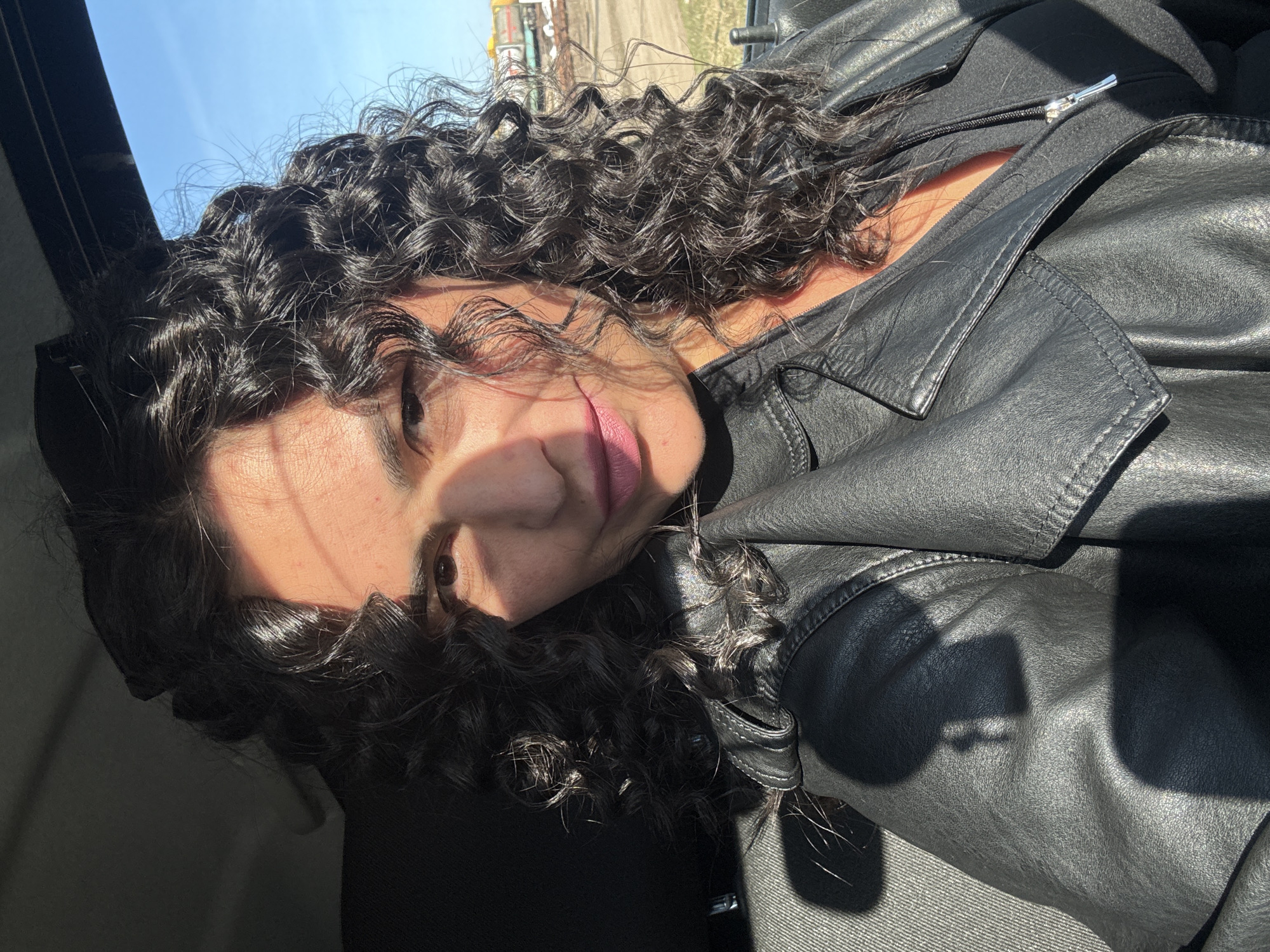 The image displays a close-up portrait of a person with dark, curly hair, taken from inside what appears to be a vehicle. The subject is wearing a black leather jacket over a black top. Partially visible facial features include an eye, part of the nose, and lips adorned with light pink lipstick.
Strong, direct sunlight illuminates portions of the person's face, their dark, shiny curls, and the textured black leather jacket, creating prominent highlights and deep shadows. A distinct shadow pattern falls across the face and jacket. Through the vehicle's window, a bright, clear blue sky is visible, along with distant, somewhat blurred landscape elements under the bright sunlight. This suggests a sunny daytime scene in Kauchuk Stantsiya, Russia. The person appears to be looking upwards, possibly leaning back in a car seat. There is no visible text in the image.