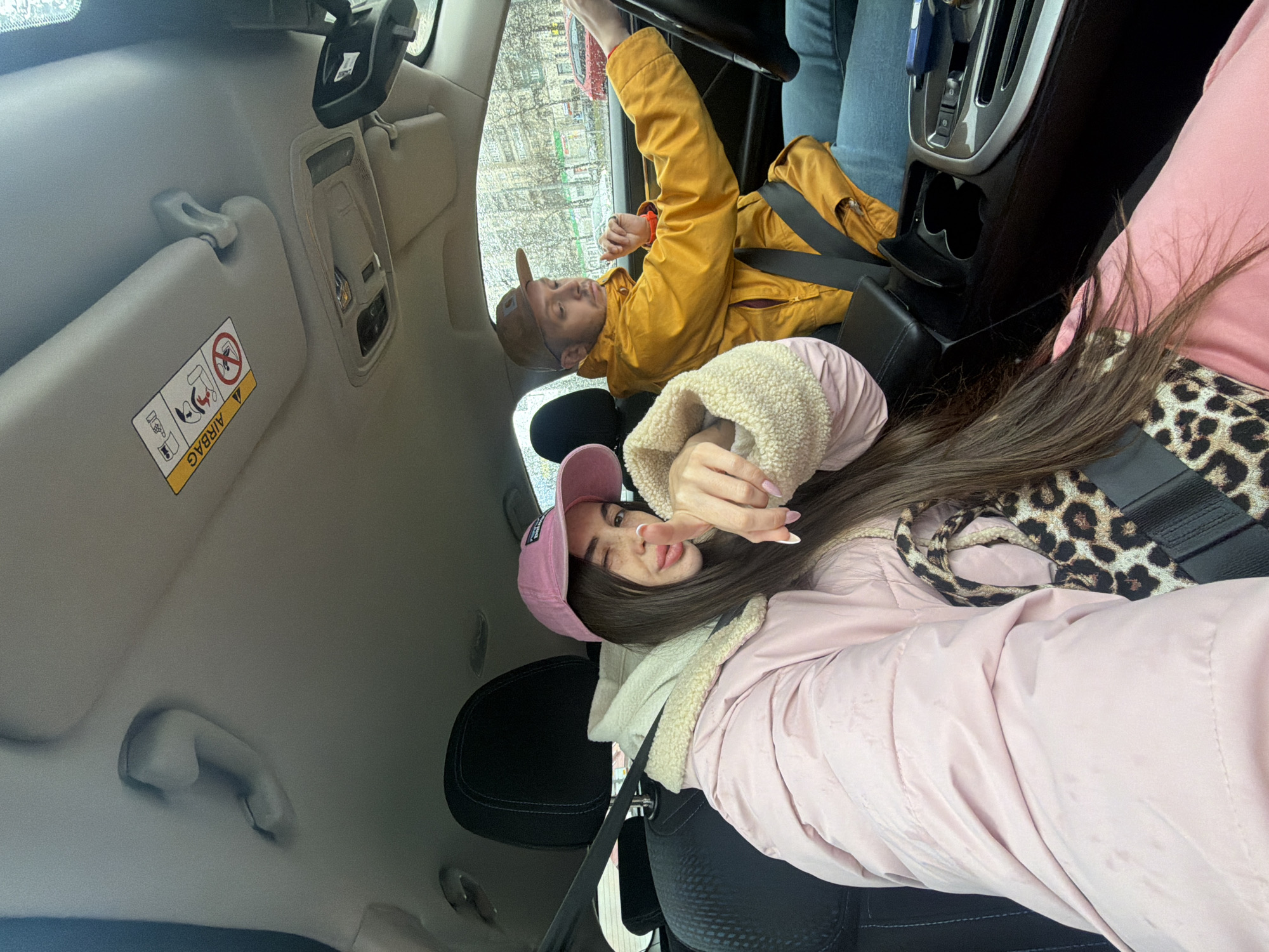 The image shows two people inside a car, presumably in Saint Petersburg, Russia, based on the architecture visible through the car window. The person in the passenger seat closest to the camera is a female wearing a pink cap, a pink jacket with a white fuzzy collar, and a leopard print scarf. Her dark hair is long. She is posing for the camera with a hand gesture. The person in the driver's seat is a male wearing a brown cap and a yellow jacket, also with a seatbelt. The car's interior is visible, including the dashboard, sun visor, and passenger-side grab handle. Outside the car, the urban environment suggests a street with buildings under overcast skies, possibly with rain or moisture on the windows.