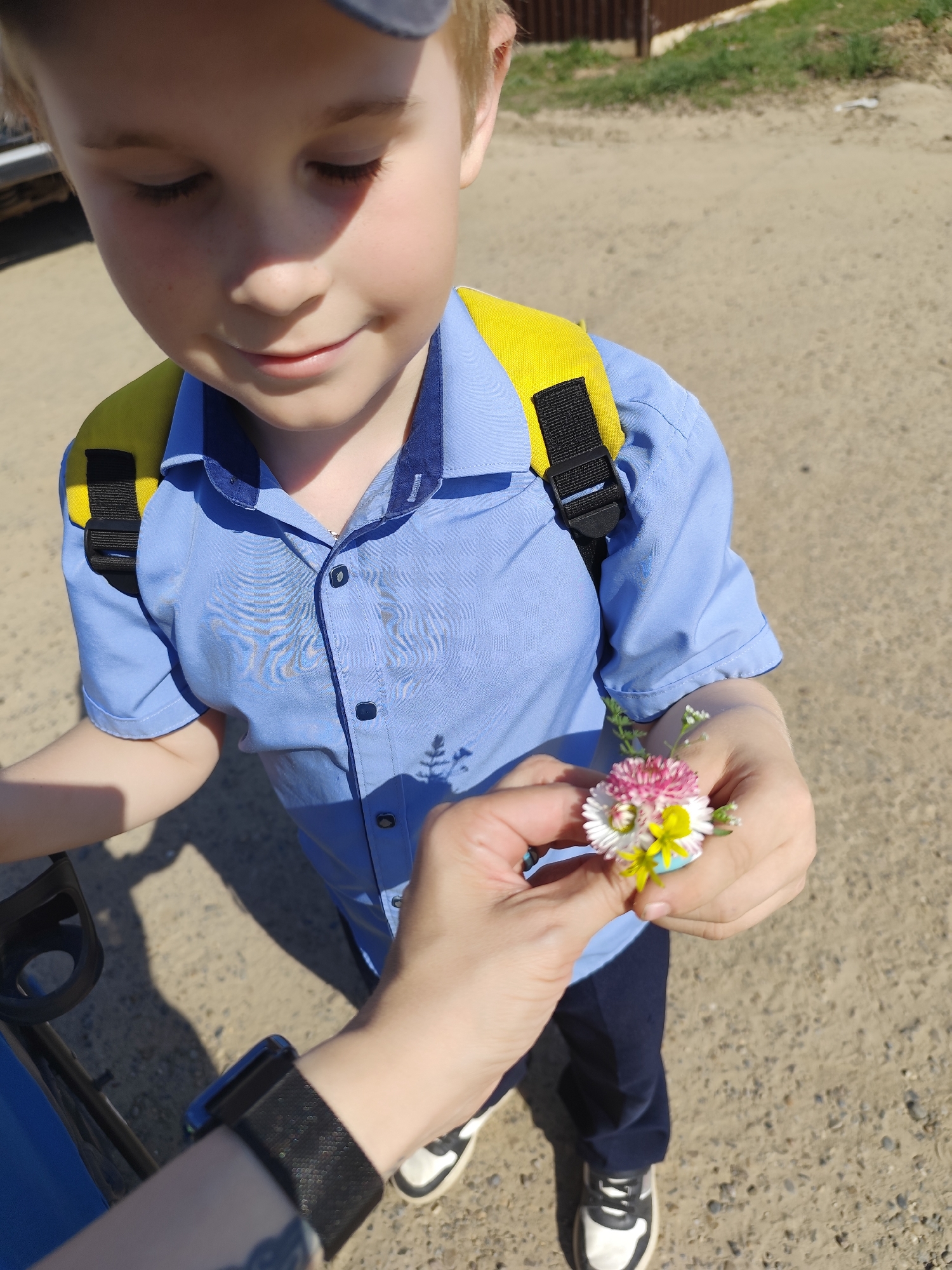 A fair-skinned boy with blond hair, wearing a blue short-sleeved collared shirt and a yellow backpack with black straps, is looking down at a small bouquet of wildflowers being held up to him by a person. The person's hand is visible, adorned with a black watch, and they are wearing black and white sneakers. The background consists of a dirt road and glimpses of a fence and vegetation. The location is Фоки, Russia.